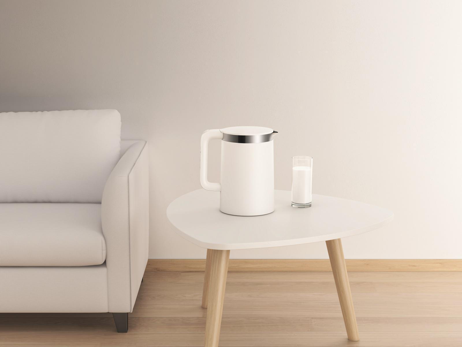 Ein weißer Wasserkocher und ein Glas Milch auf einem weißen Tisch neben einem weißen Sofa.