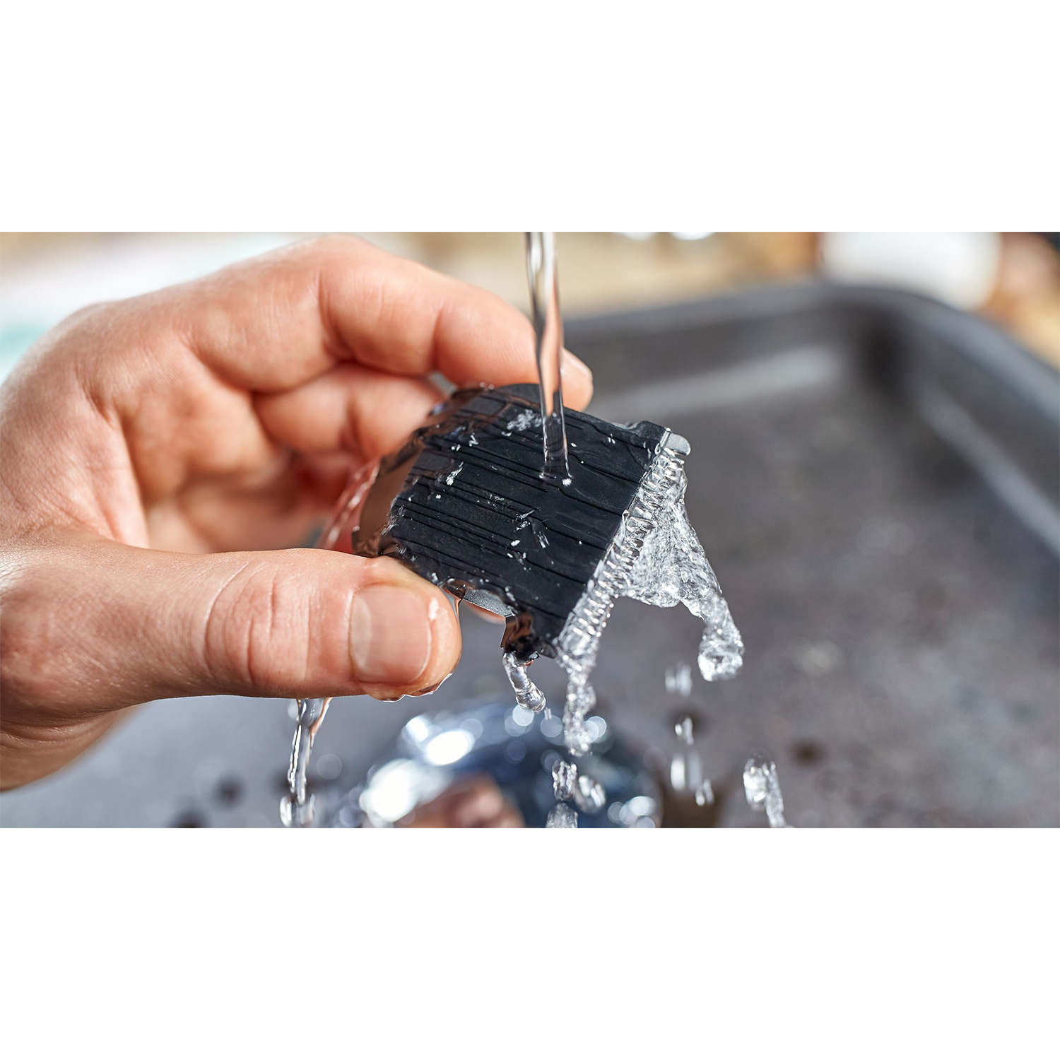 Mano che risciacqua una testina di rasoio elettrico nera sotto l'acqua corrente. Vassoio d'argento sullo sfondo.