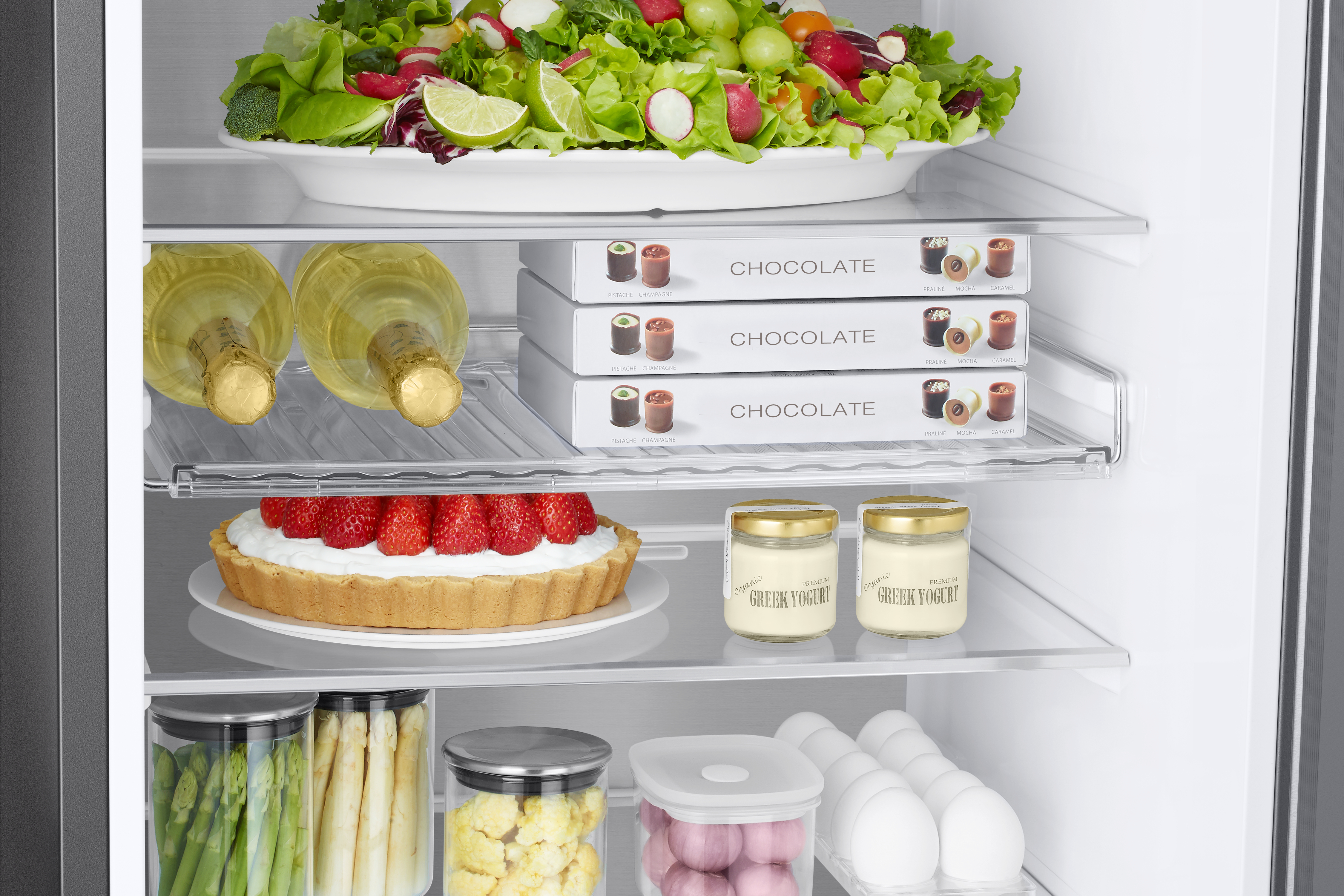 Kühlschrankinneres mit Salat, Schokolade, Joghurt und einer Torte. Lebensmittel sind auf Regalen angeordnet.