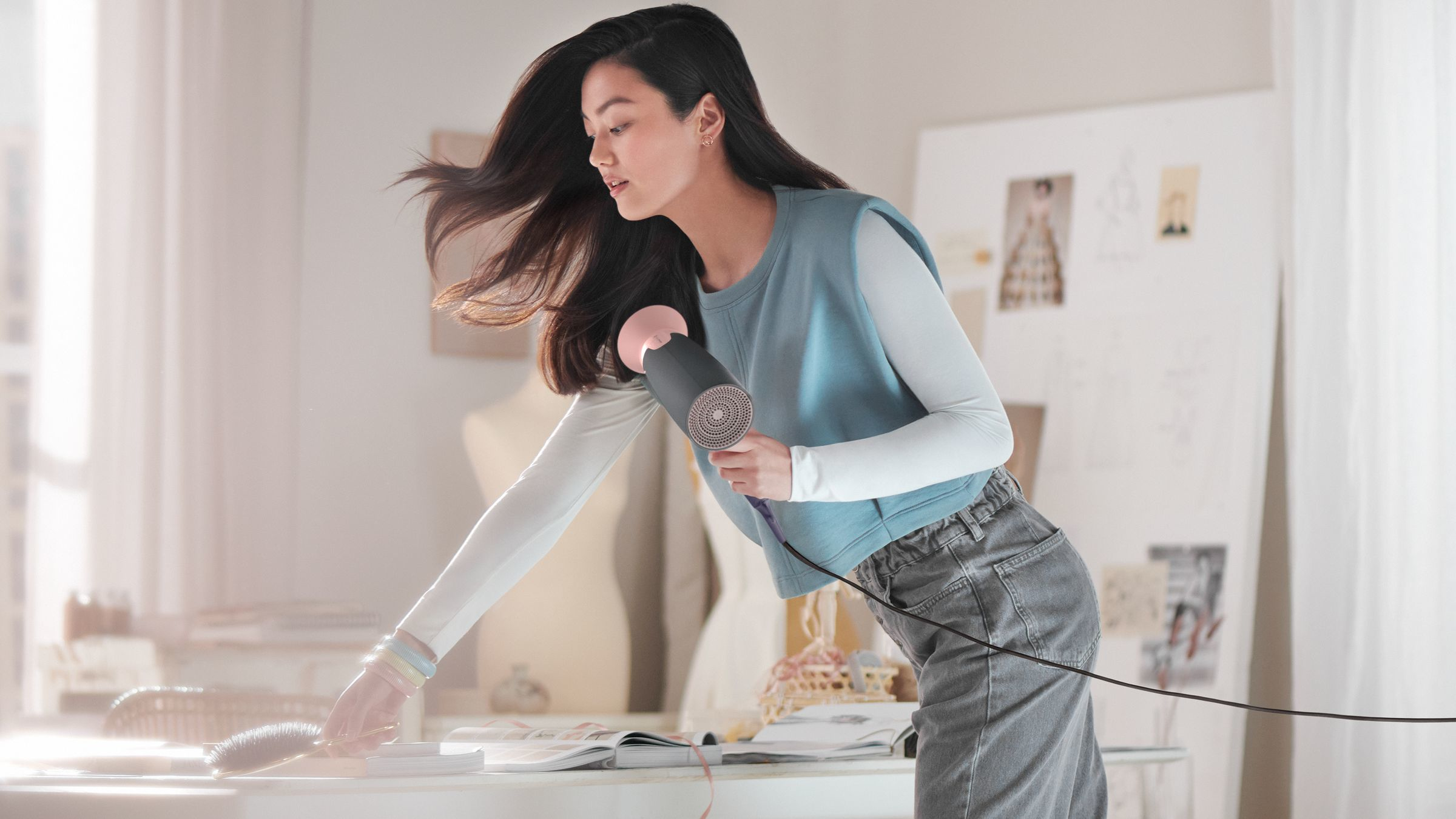 Une femme se sèche les cheveux avec un sèche-cheveux sur une table blanche avec un livre ouvert et un mannequin.