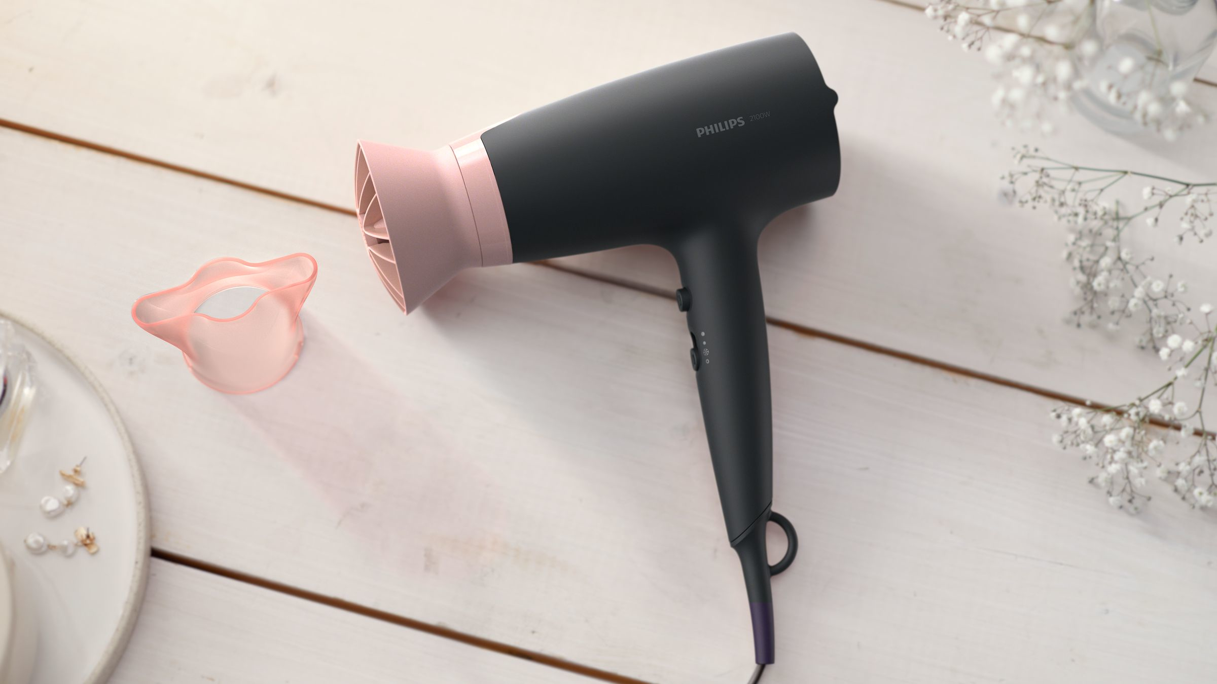 Sèche-cheveux noir et rose sur une surface en bois blanc avec un accessoire rose et des fleurs blanches.