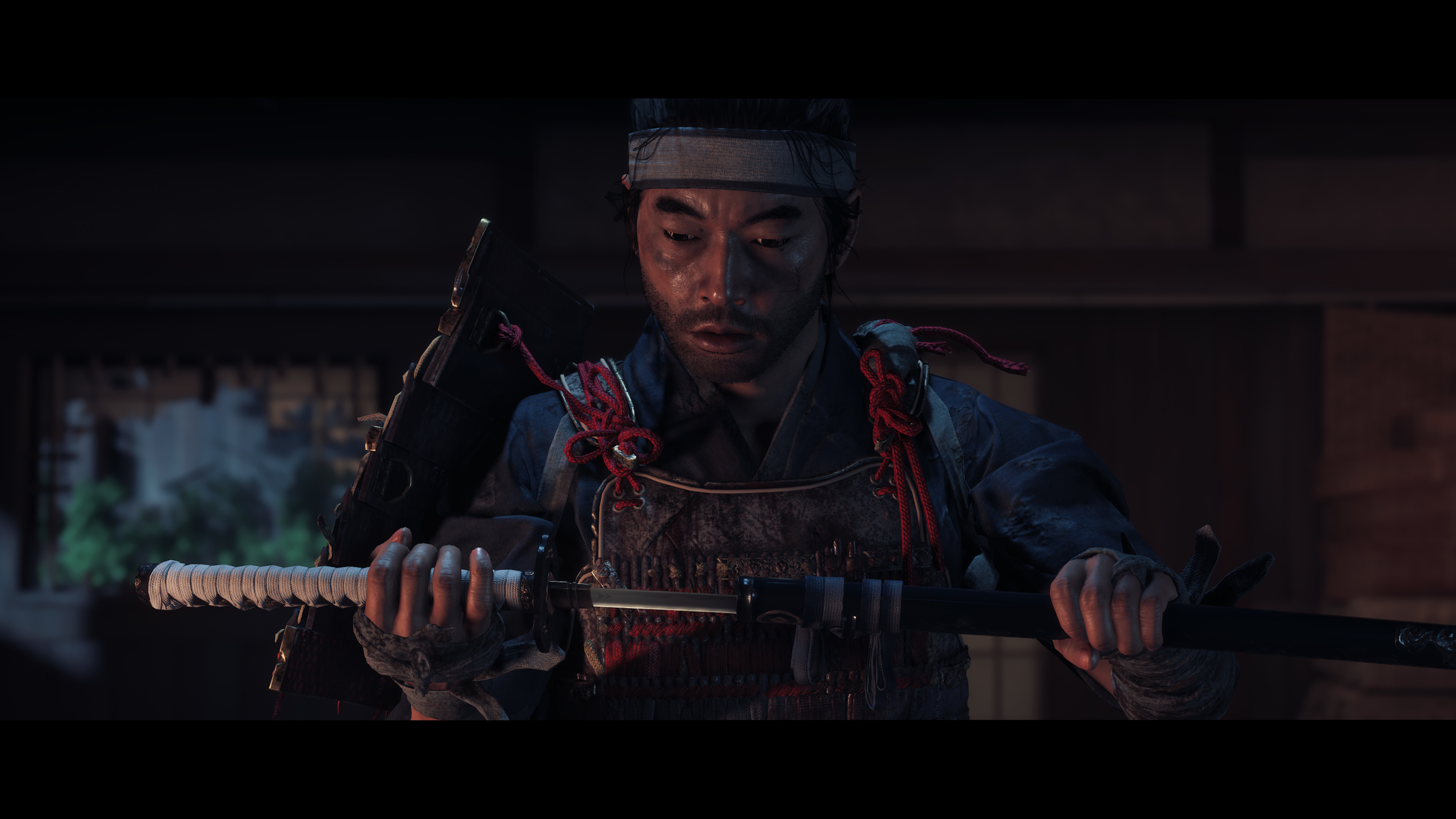 Uomo in abito da samurai che tiene una spada, guardando in basso in una stanza poco illuminata.