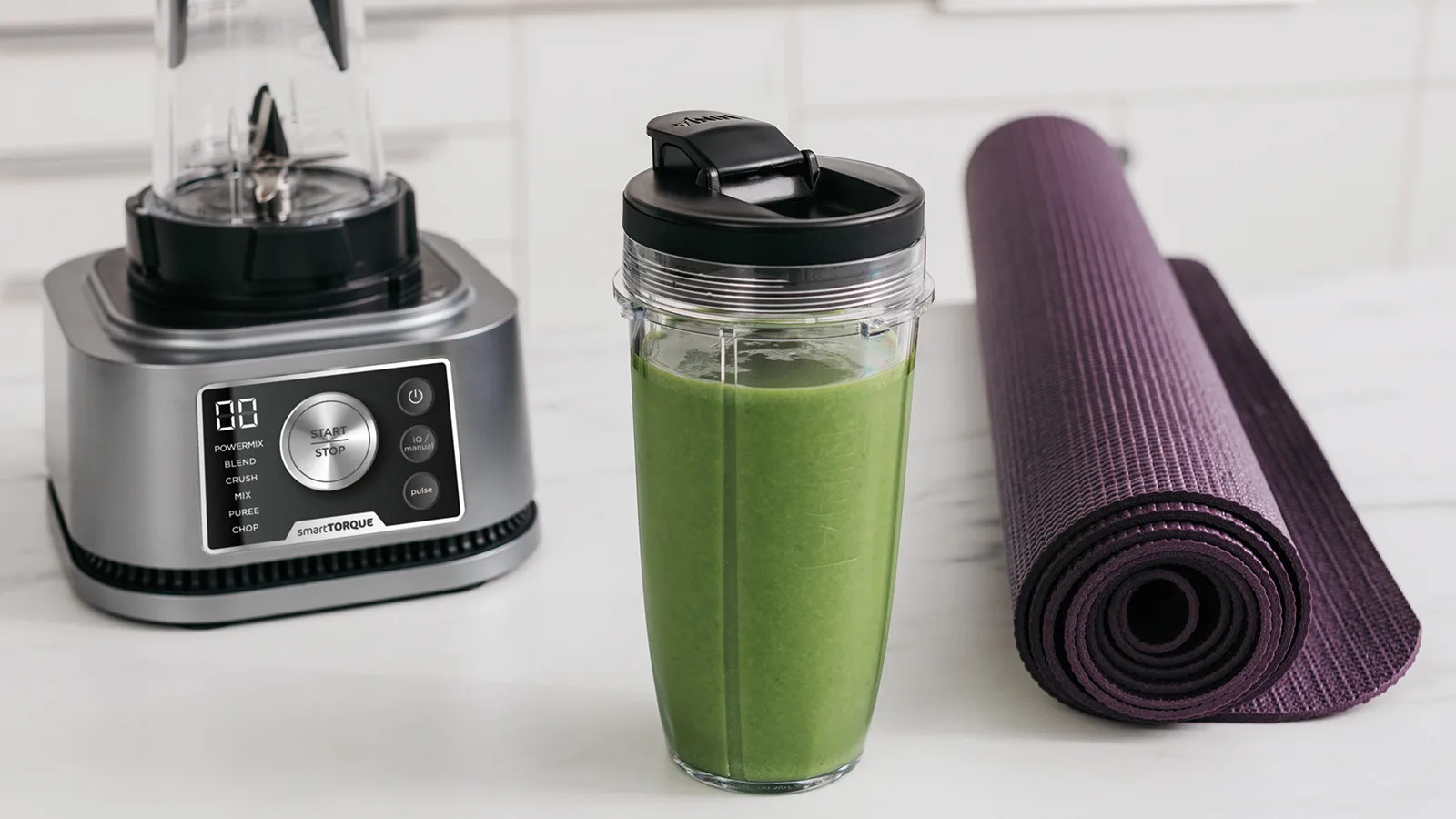 Ein Mixer, ein Smoothie-Becher und eine zusammengerollte lila Yogamatte stehen auf einer weißen Arbeitsplatte.