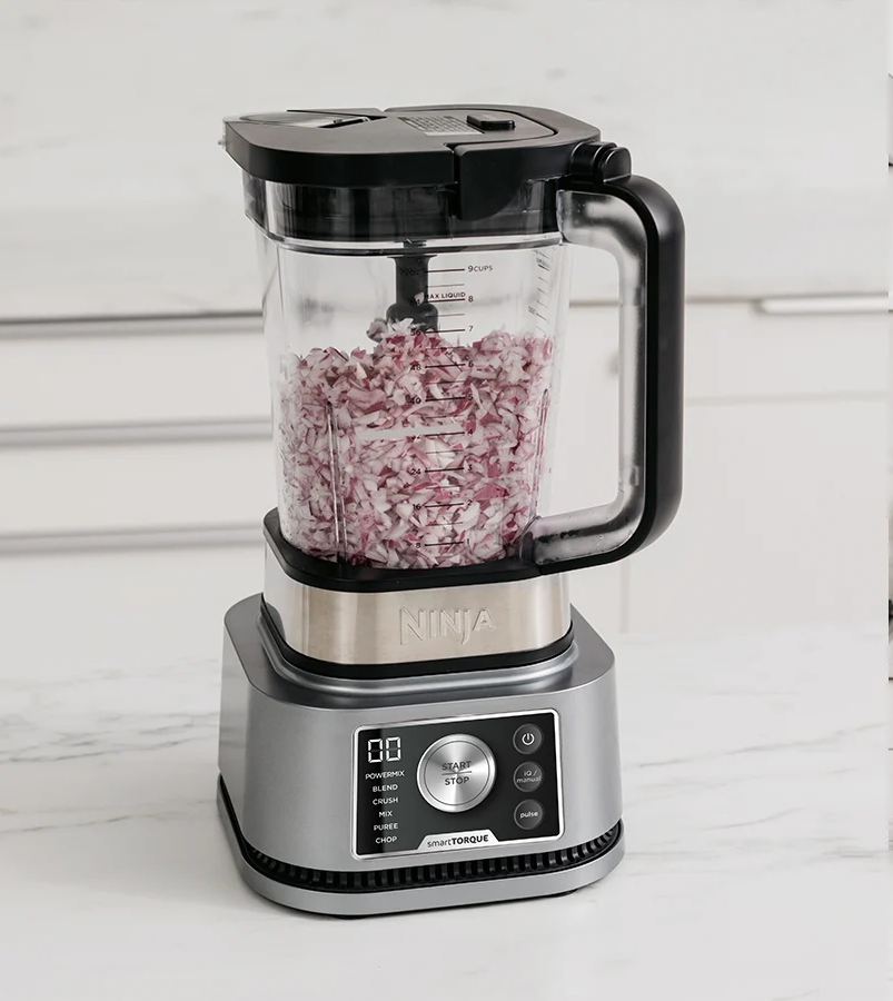 Ein Ninja-Mixer mit gehackten roten Zwiebeln. Der Mixer ist silberfarben mit schwarzem Deckel und Griff. Eine Küchenarbeitsplatte ist der Hintergrund.
