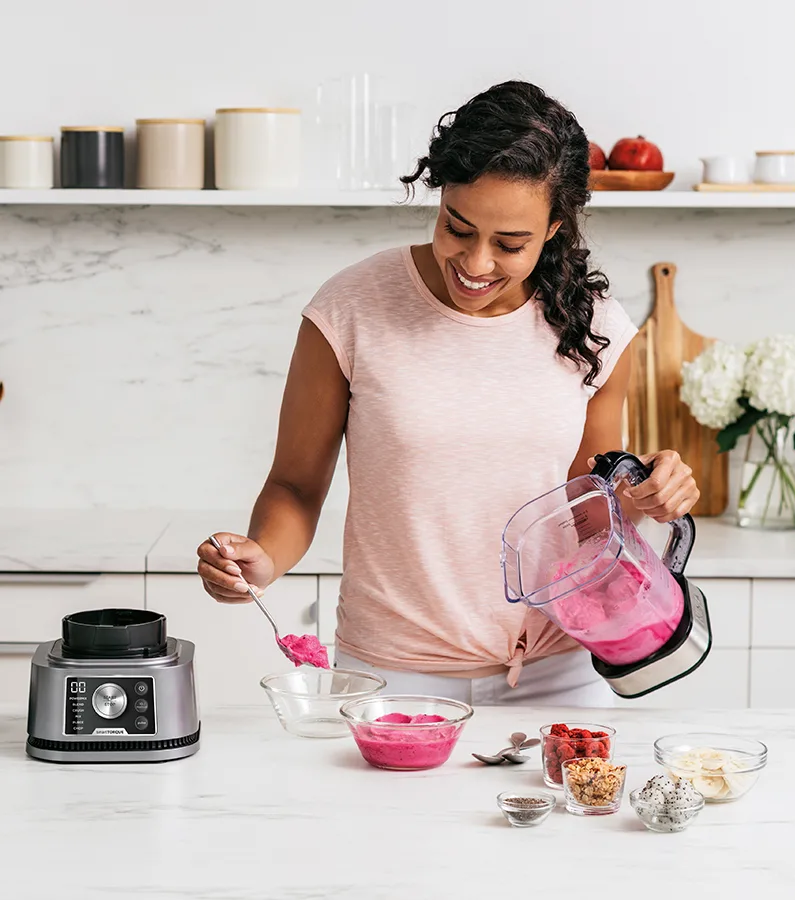 Frau macht Smoothie. Sie gießt es aus einem Mixer in Schüsseln. Zutaten wie Beeren und Drachenfrucht sind auf der Theke.