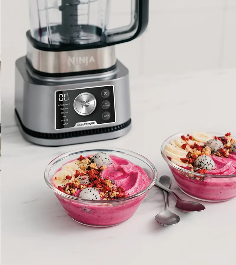 Ein Ninja-Mixer steht über zwei Schüsseln mit rosa Smoothie, belegt mit Bananen, Drachenfrucht und Müsli, mit Löffeln.
