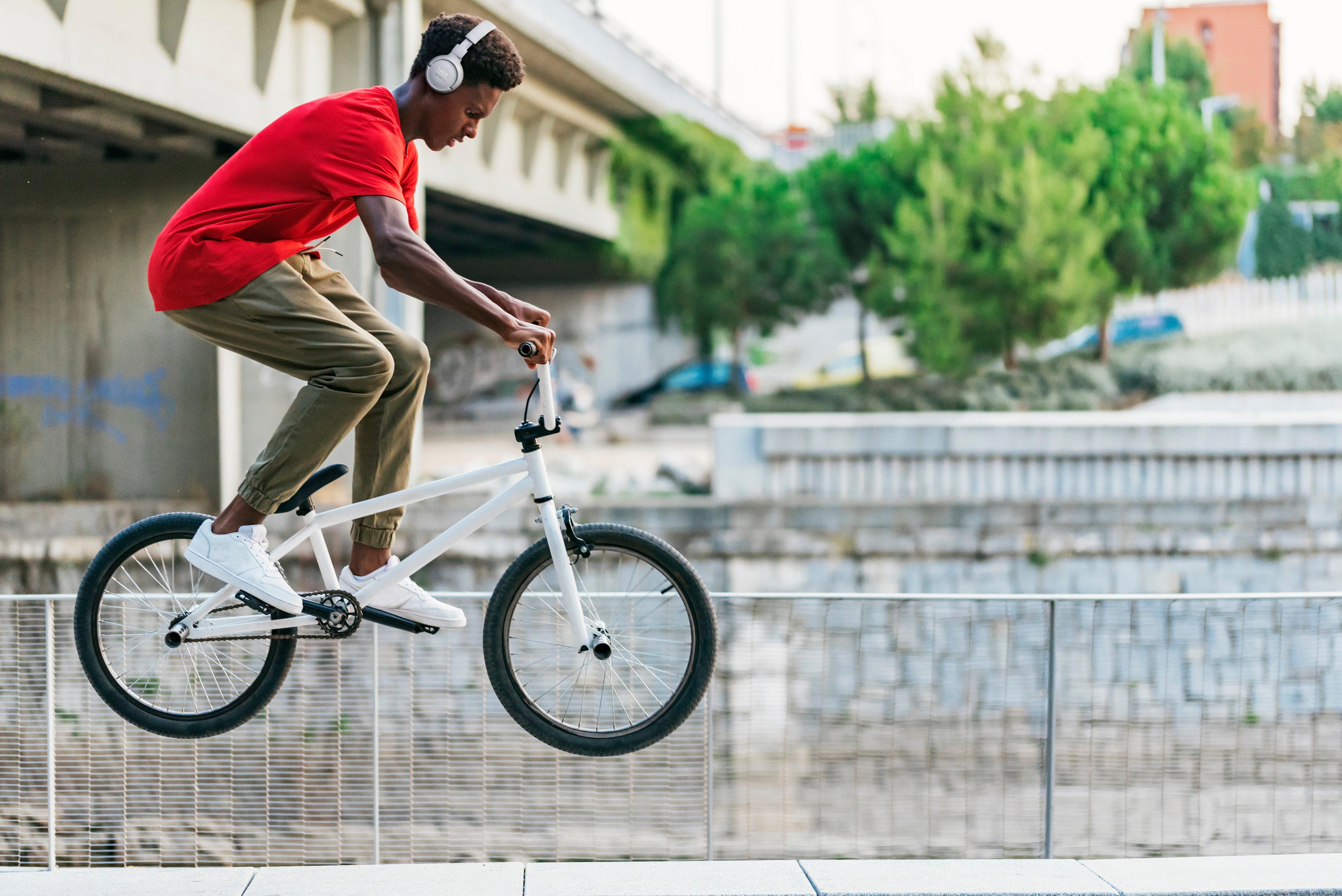 Kırmızı gömlek giyen, beyaz BMX bisiklet üzerinde havada genç bir adam. Arka planda bir köprü ve yeşillik.