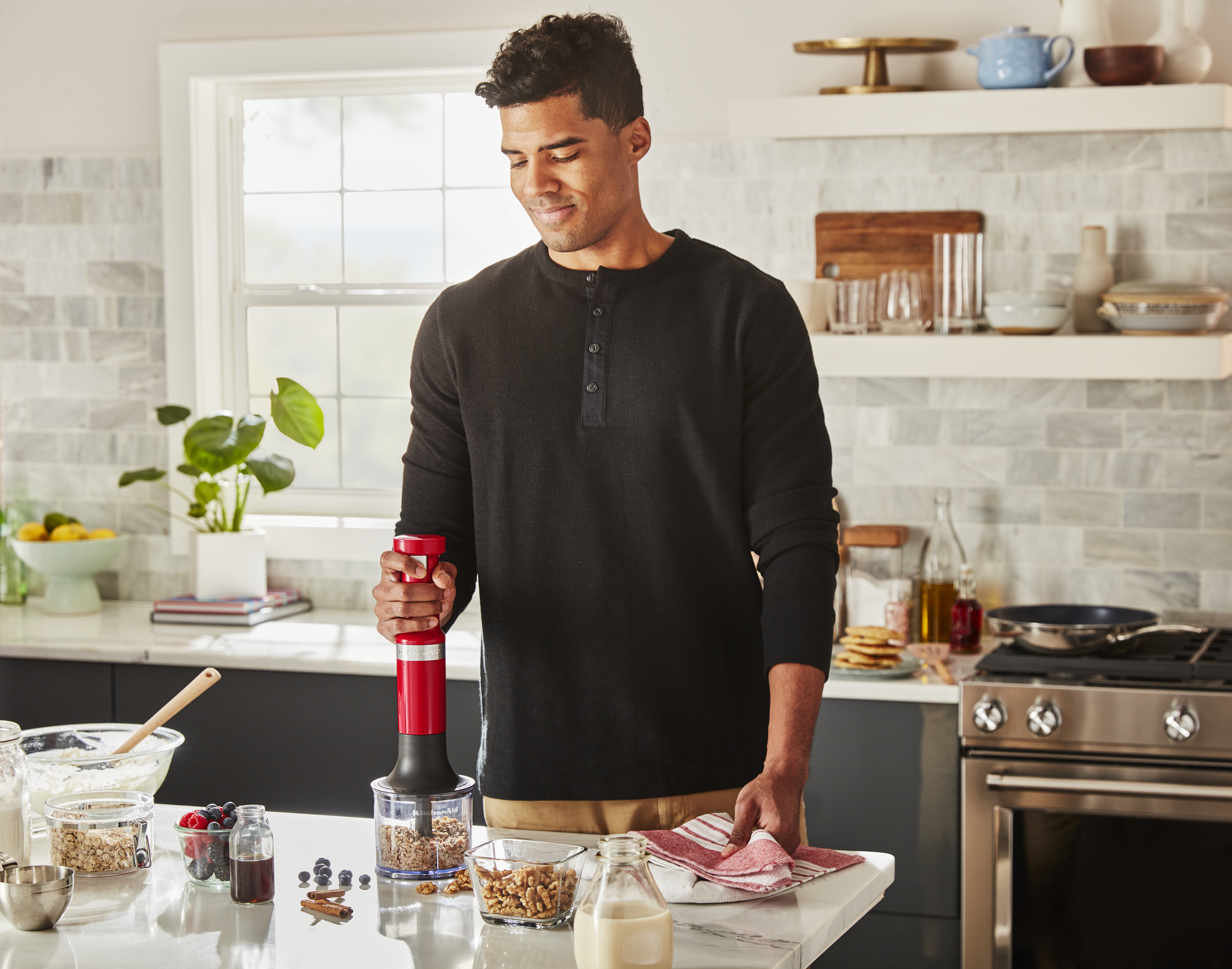 Mann, der einen roten Stabmixer in einer Schüssel auf einer Küchentheke verwendet. Beeren und Nüsse sichtbar.