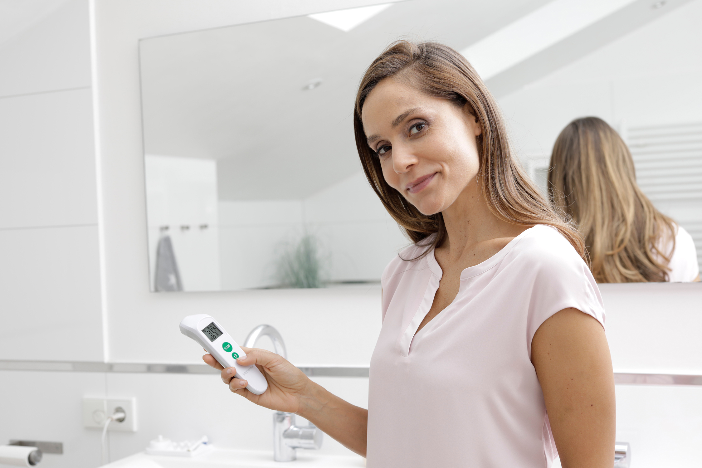 Een vrouw in een roze shirt die een thermometer vasthoudt in een badkamer, kijkt naar de camera.