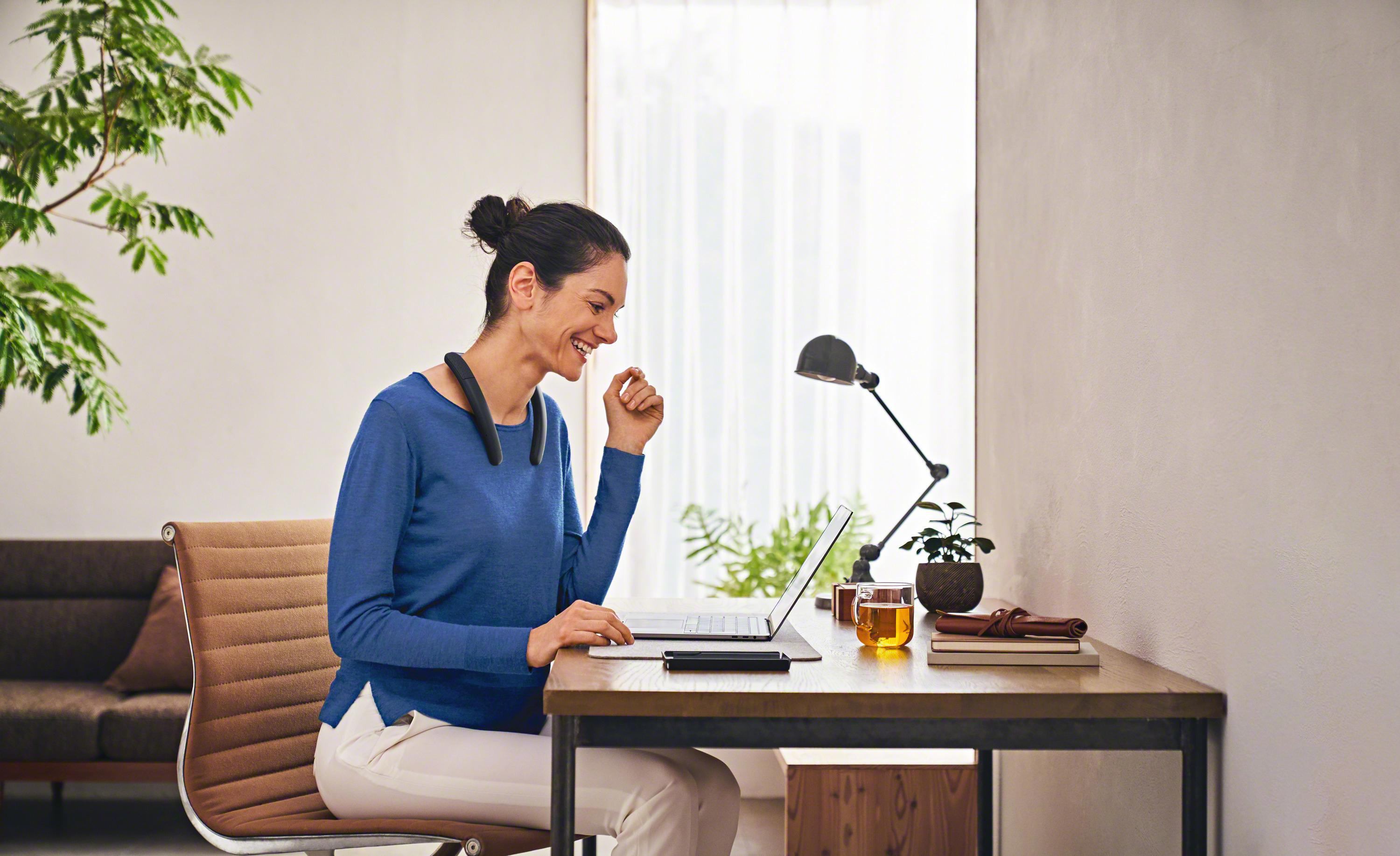 Frau arbeitet am Laptop am Schreibtisch, trägt einen Nackenlautsprecher. Tasse und Pflanze sichtbar.