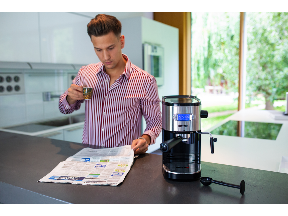 Un hombre leyendo un periódico, bebiendo de una taza, junto a una máquina de espresso Koenic.