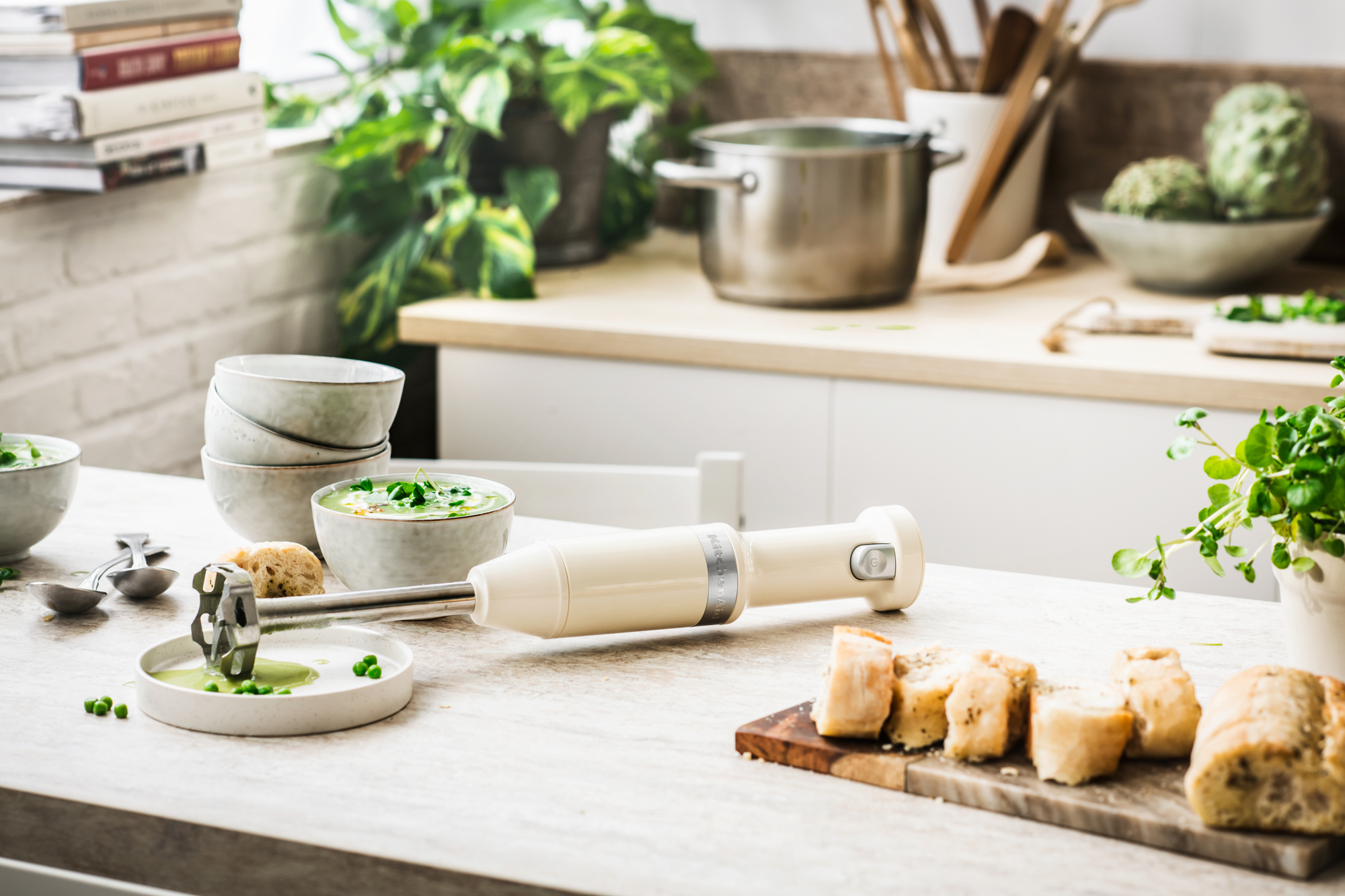 Eine Küchenszene mit einem beigen Handmixer auf einer Arbeitsplatte. Es gibt Suppenschalen und Brot.