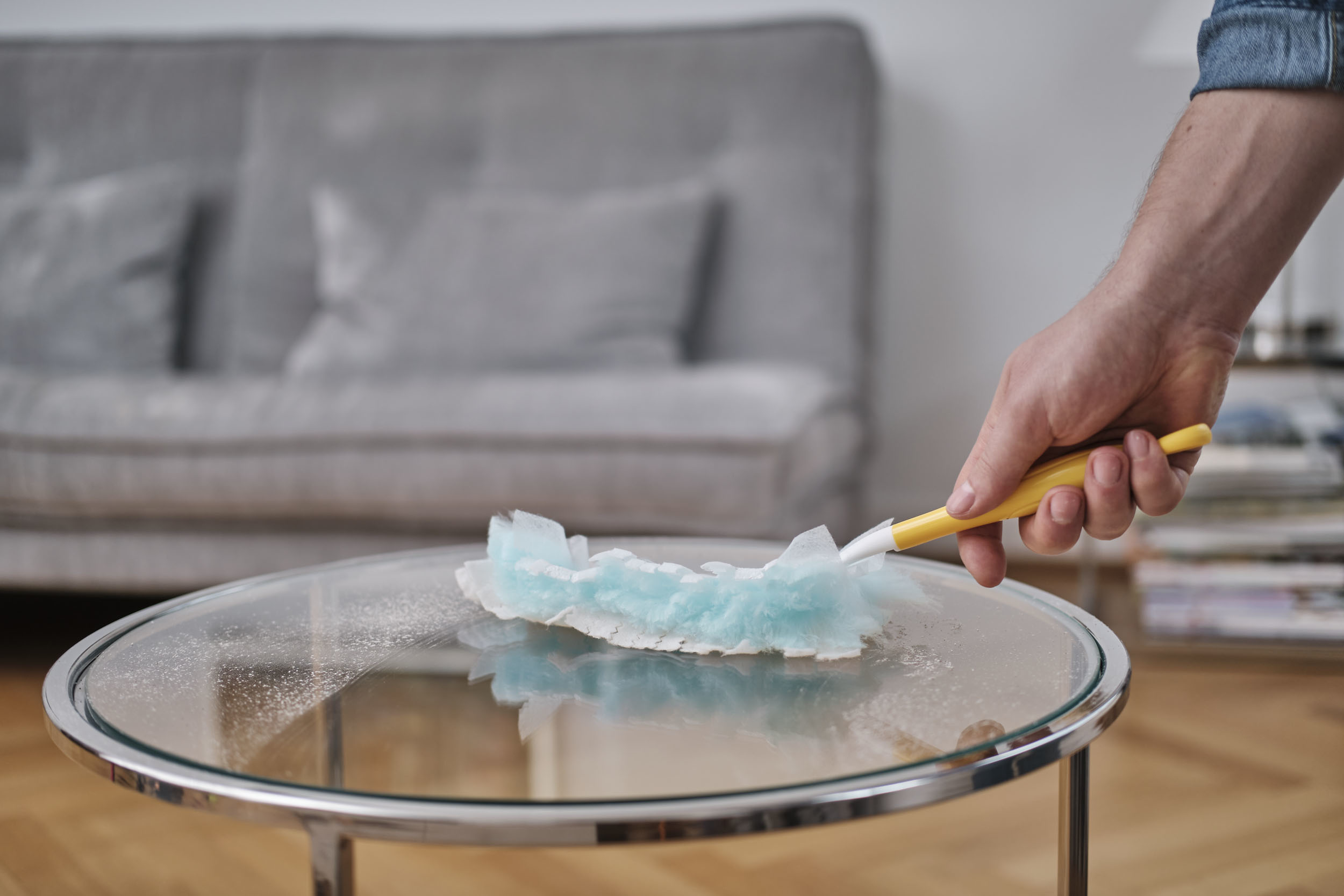 Person reinigt einen Glastisch mit einem Staubwedel, ein Sofa im Hintergrund.