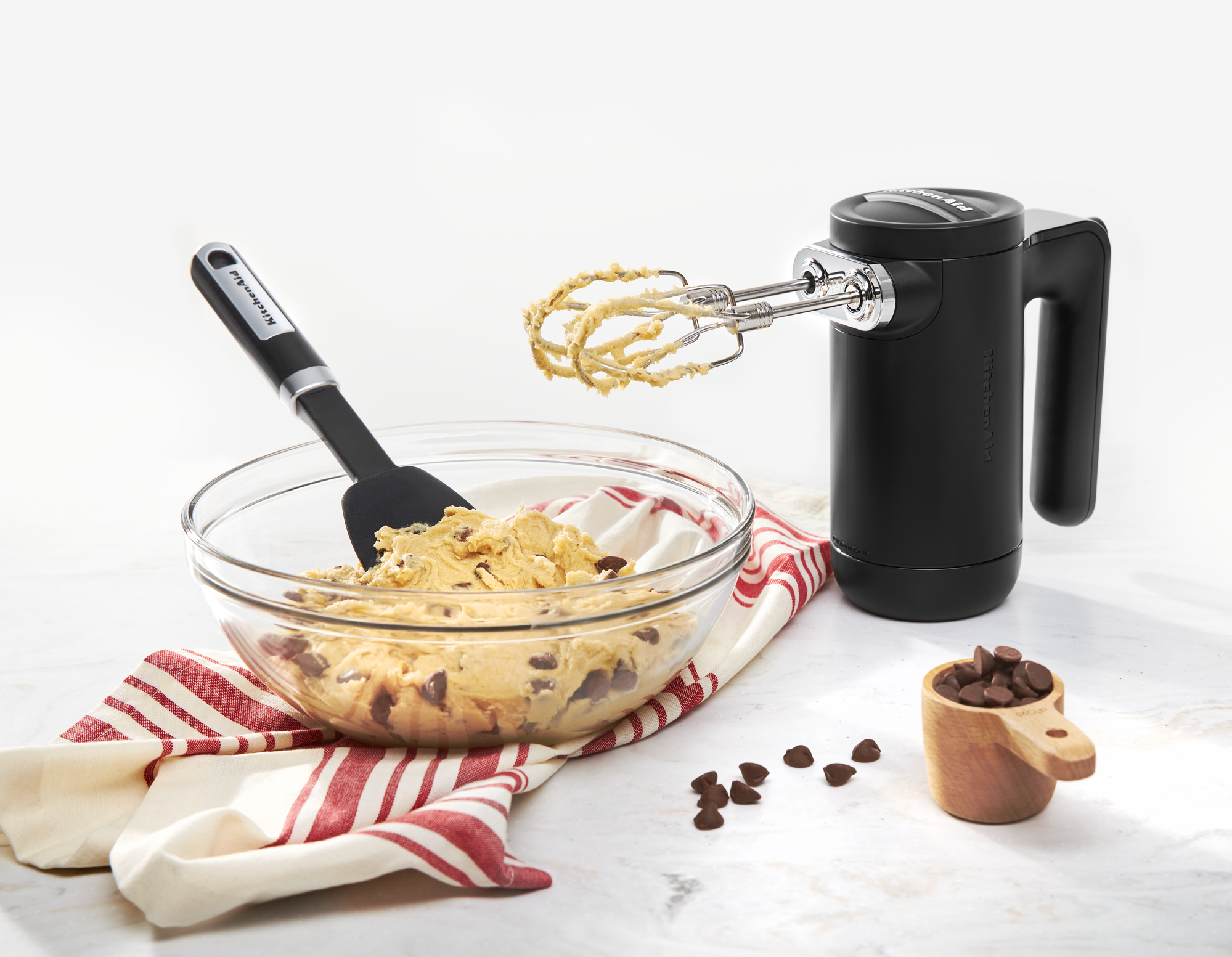 Ein KitchenAid Handmixer mischt Keksteig in einer Glasschüssel, mit Spatel und Schokoladenstückchen.