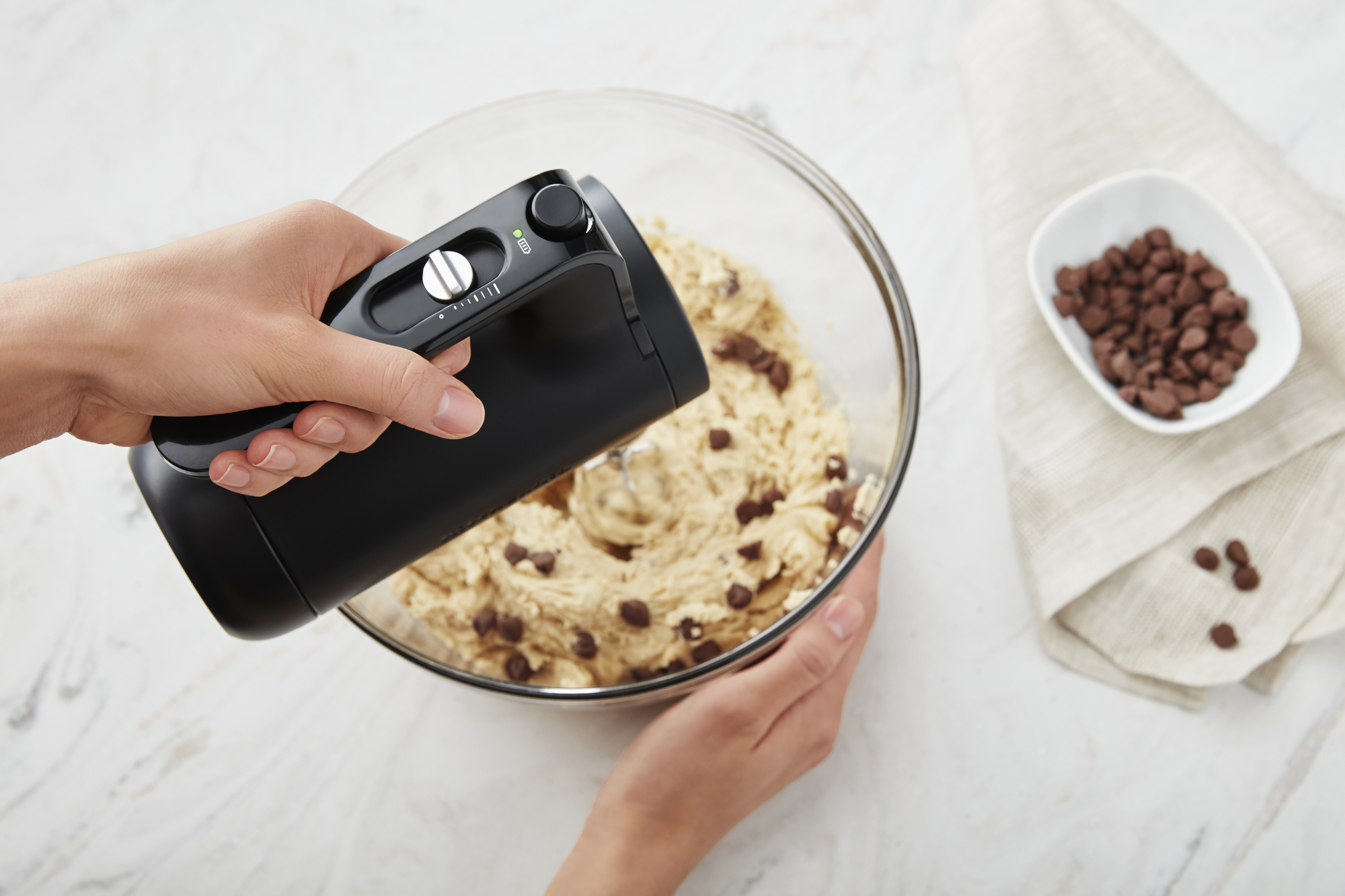 Hände halten einen schwarzen Handmixer über einer Schüssel mit Keksteig mit Schokostückchen.