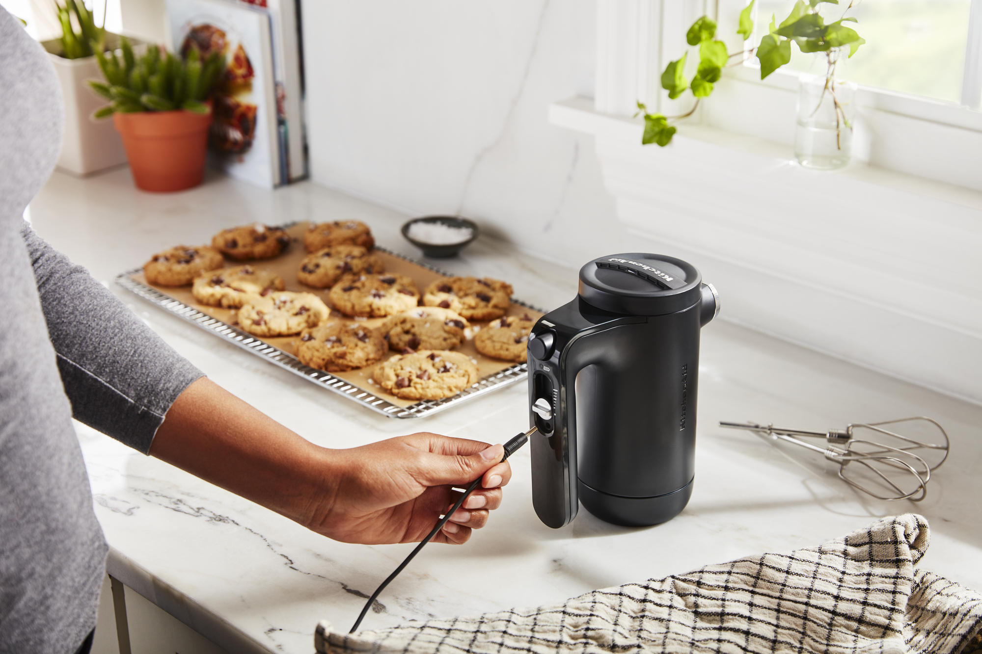 Eine Person steckt ein Ladekabel in einen schwarzen KitchenAid Handmixer, mit Keksen auf einem Tablett in der Nähe.
