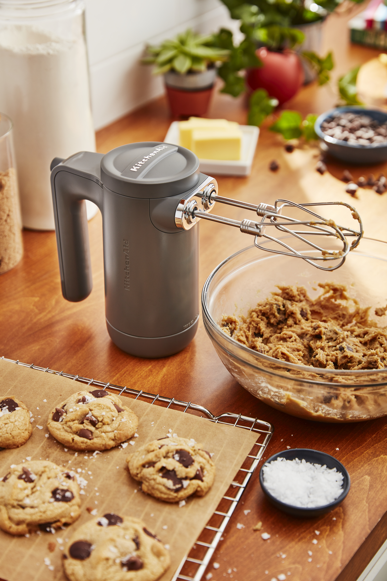 KitchenAid Handmixer, der Keksteig in einer Glasschüssel verrührt. Kekse auf Backpapier im Vordergrund.