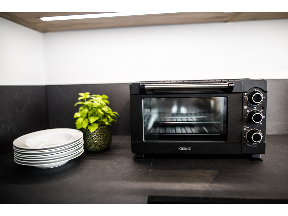 Horno tostador KOENIC negro con platos blancos y una planta. Sobre encimera negra con pared blanca y gris.