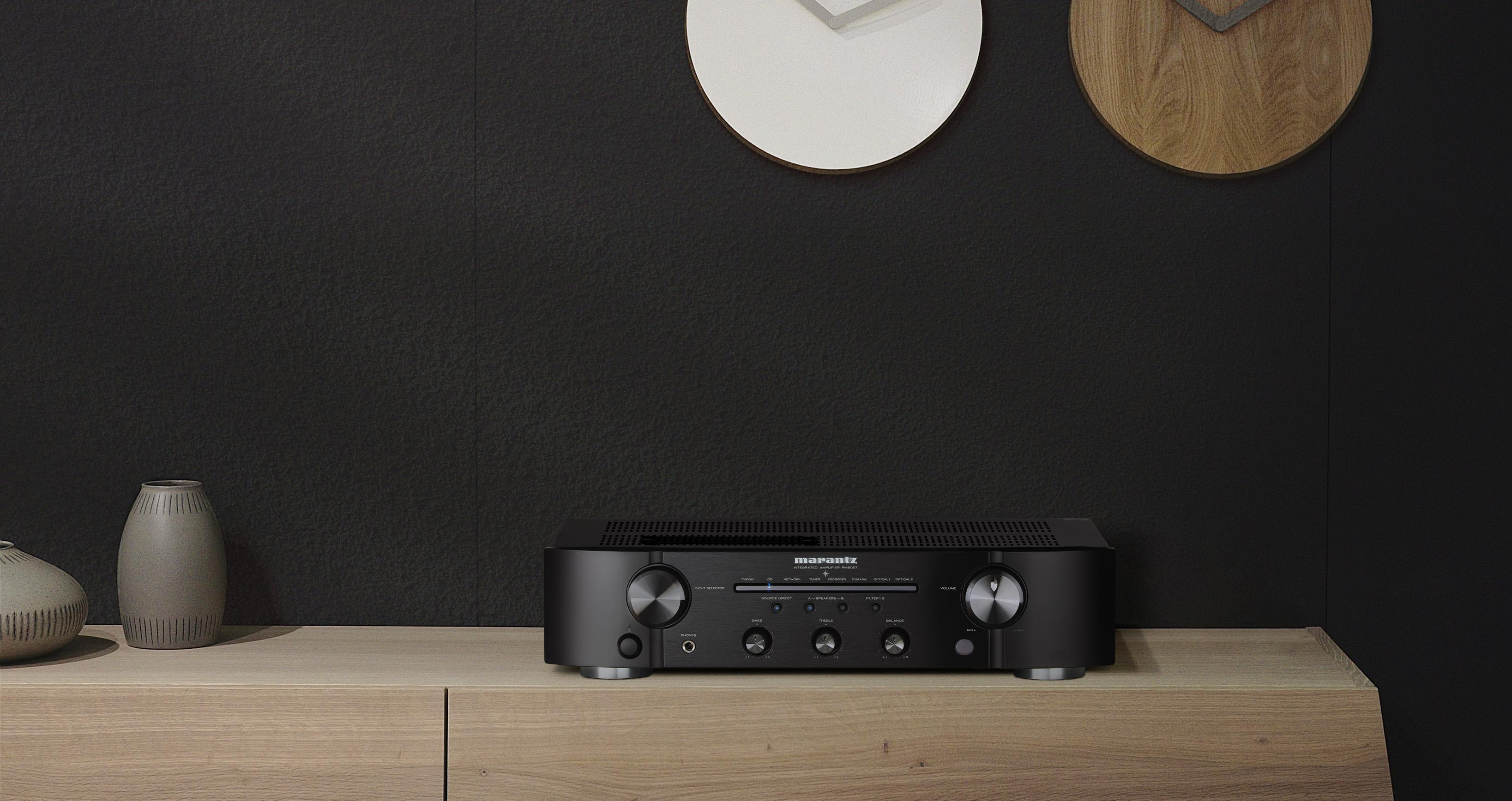 Schwarzer Audio-Receiver auf einem Holzregal. Zwei Uhren an der Wand. Dunkler Hintergrund und neutrale Farben.