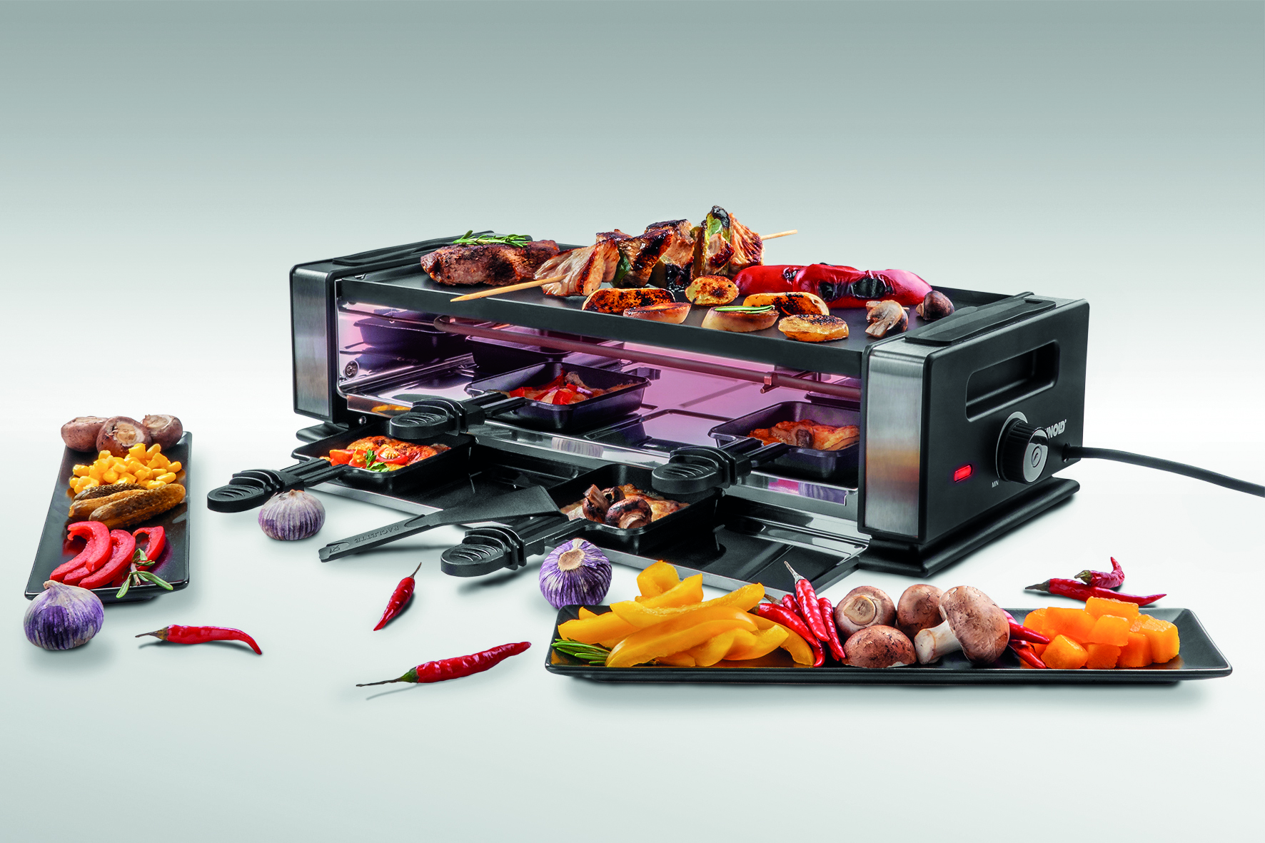 Ein Raclette-Grill mit Essen. Enthält Gemüse, Fleisch und Käse. Weißer Hintergrund.