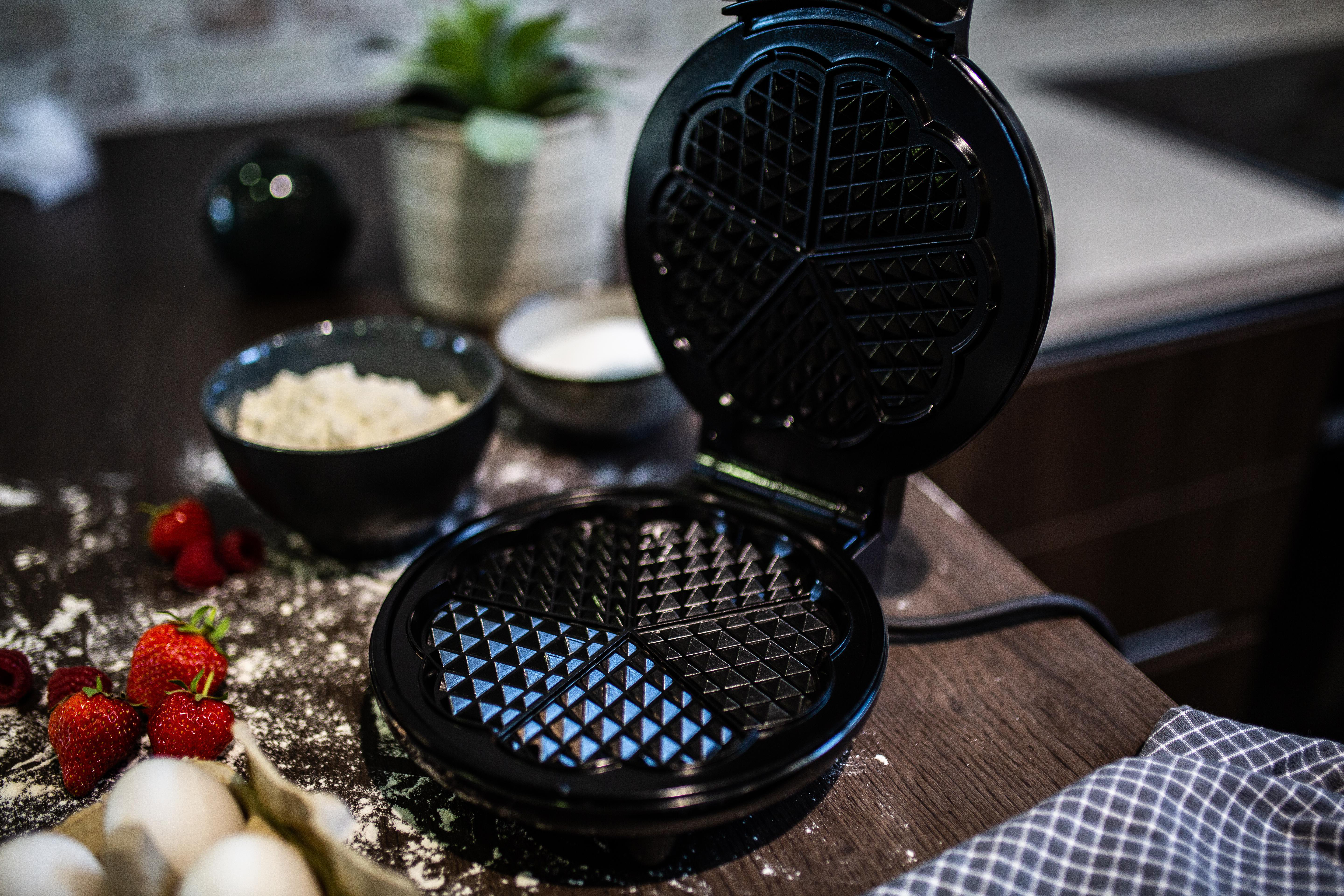 Offener schwarzer Waffeleisen mit gekochten Waffeln. Zutaten wie Eier und Erdbeeren daneben.