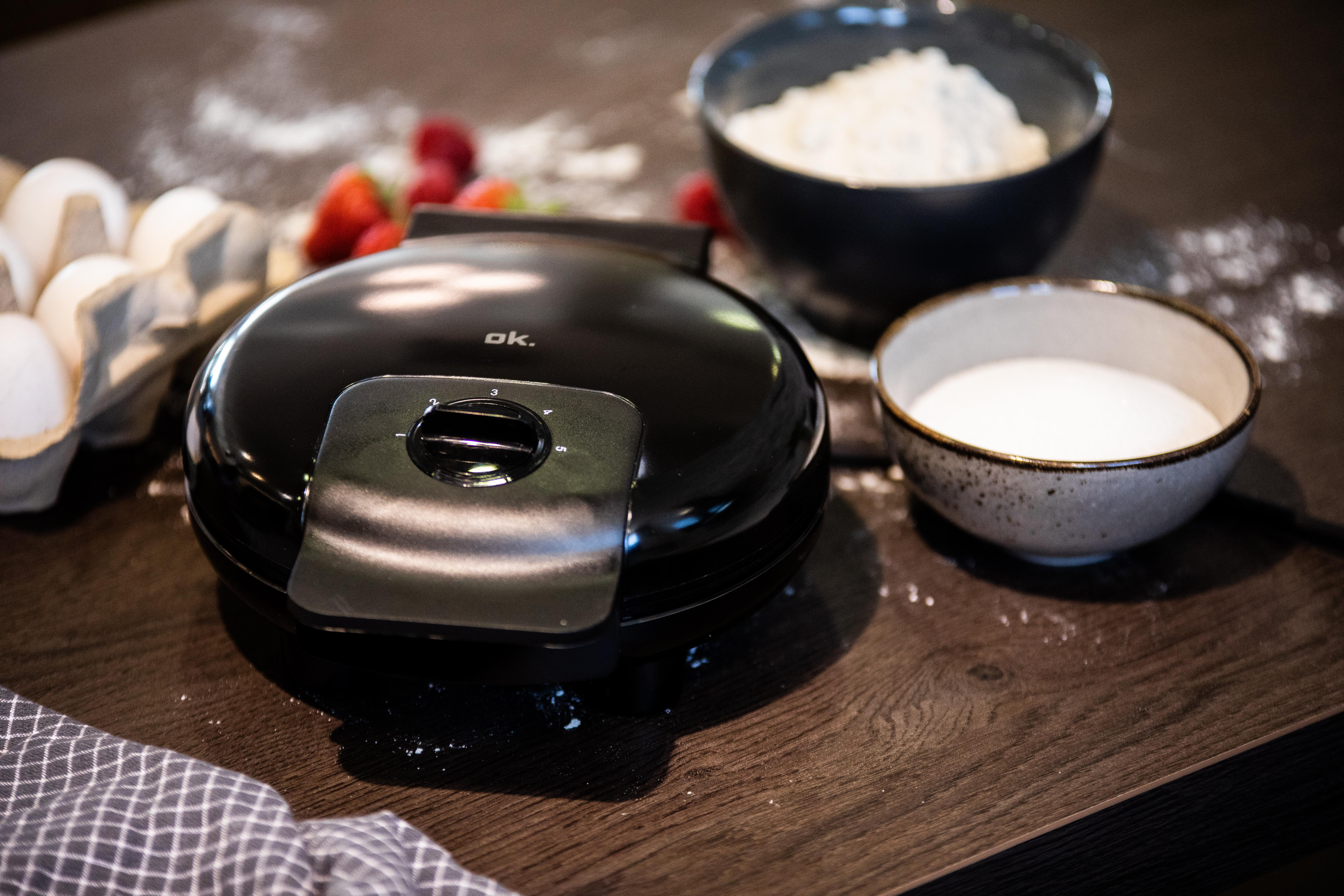 Schwarzer Waffeleisen auf Holzoberfläche. Zutaten, Schalen mit Zucker und Mehl sowie Eier sind vorhanden.