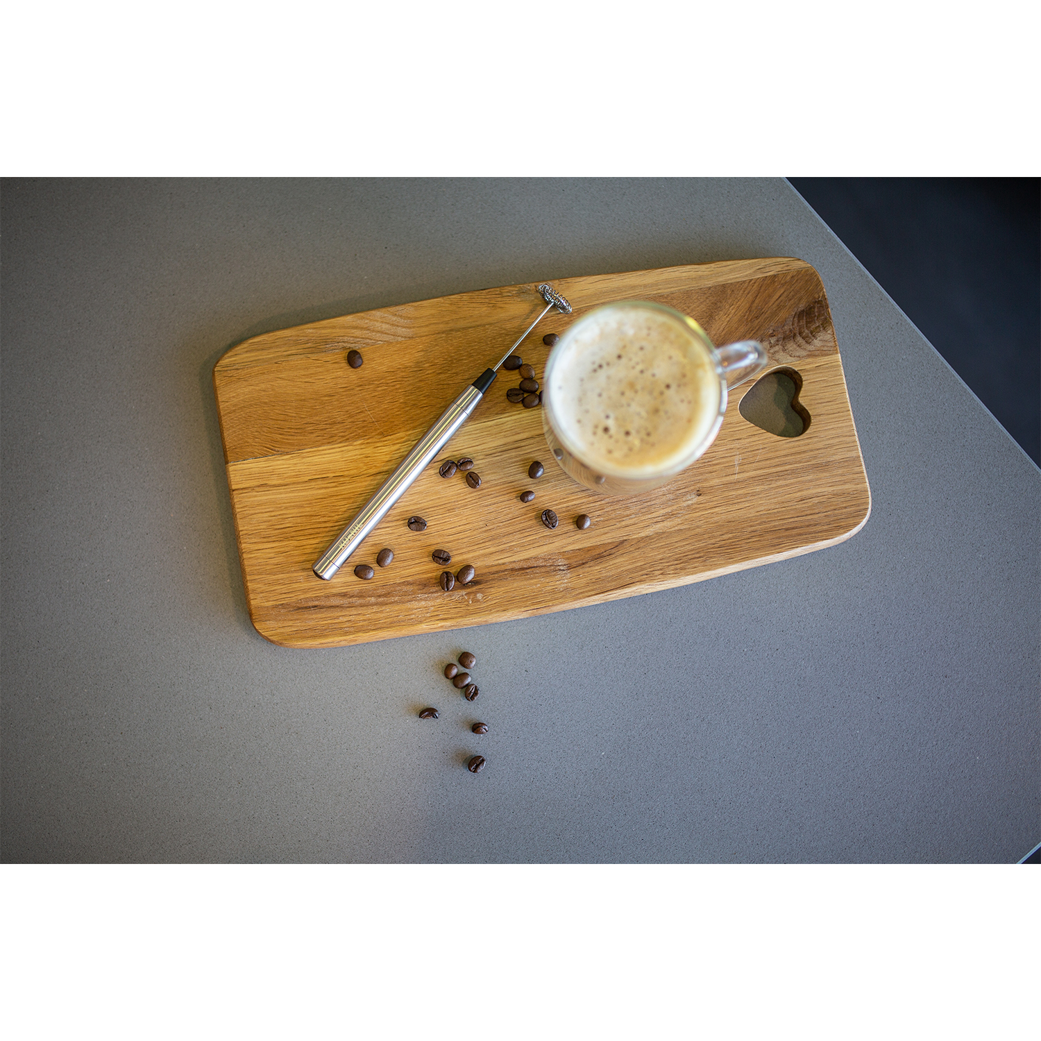 Un tagliere di legno con caffè, un montalatte e chicchi su una superficie grigia.
