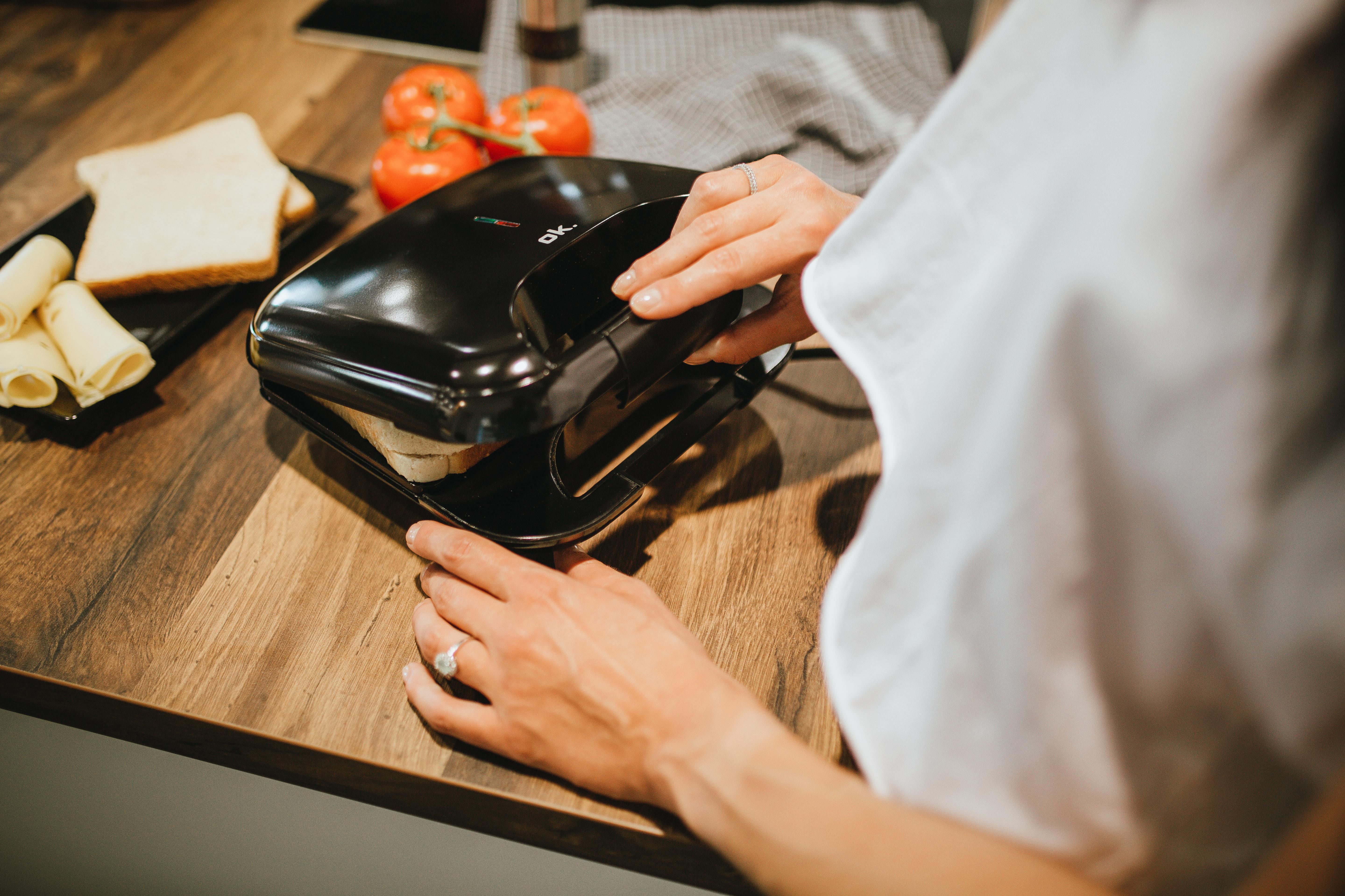 Eine Person benutzt einen Sandwichmaker. Brot und Käse liegen daneben. Tomaten sind im Hintergrund.