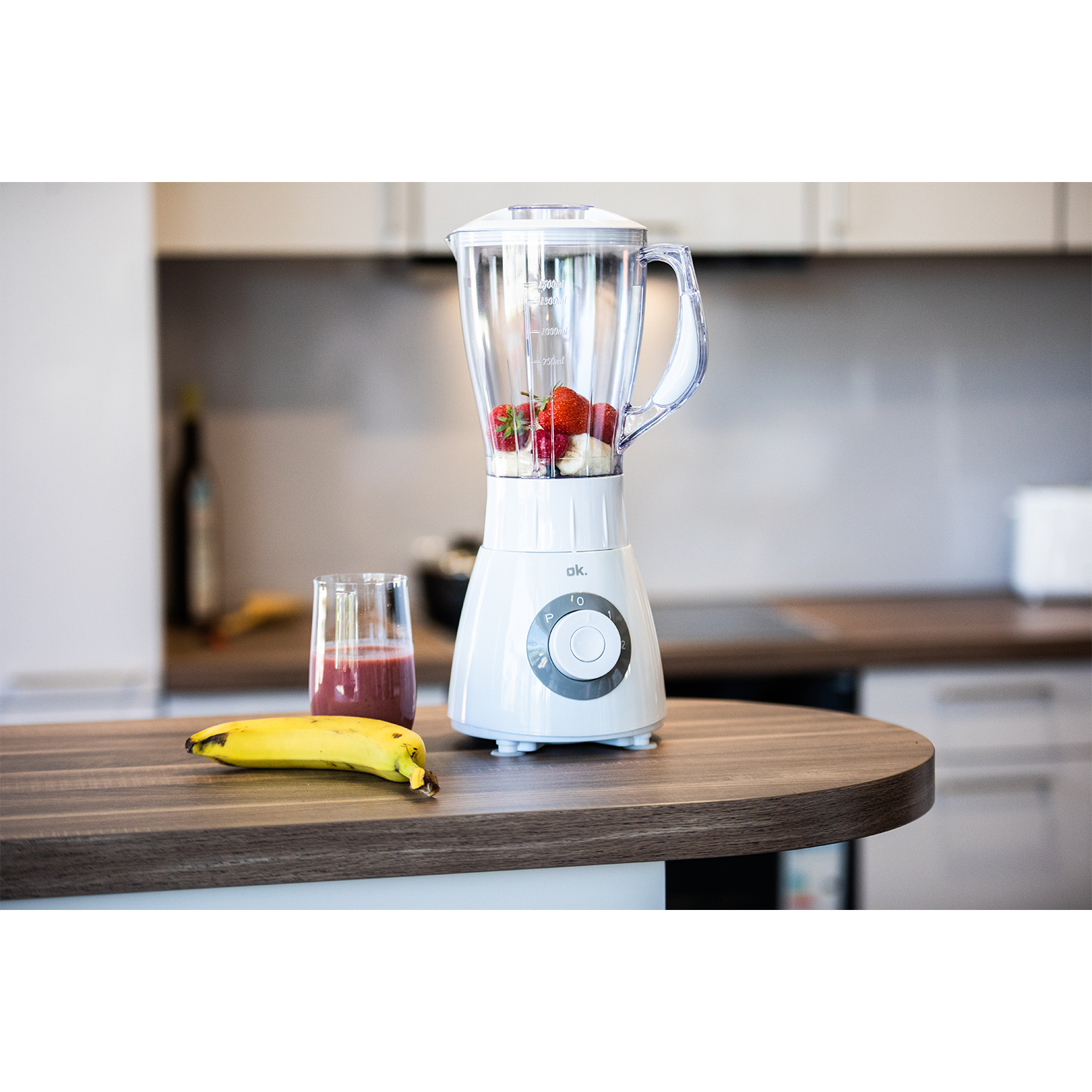Un frullatore bianco con frutta all'interno, un bicchiere di frullato e una banana sul piano di lavoro.