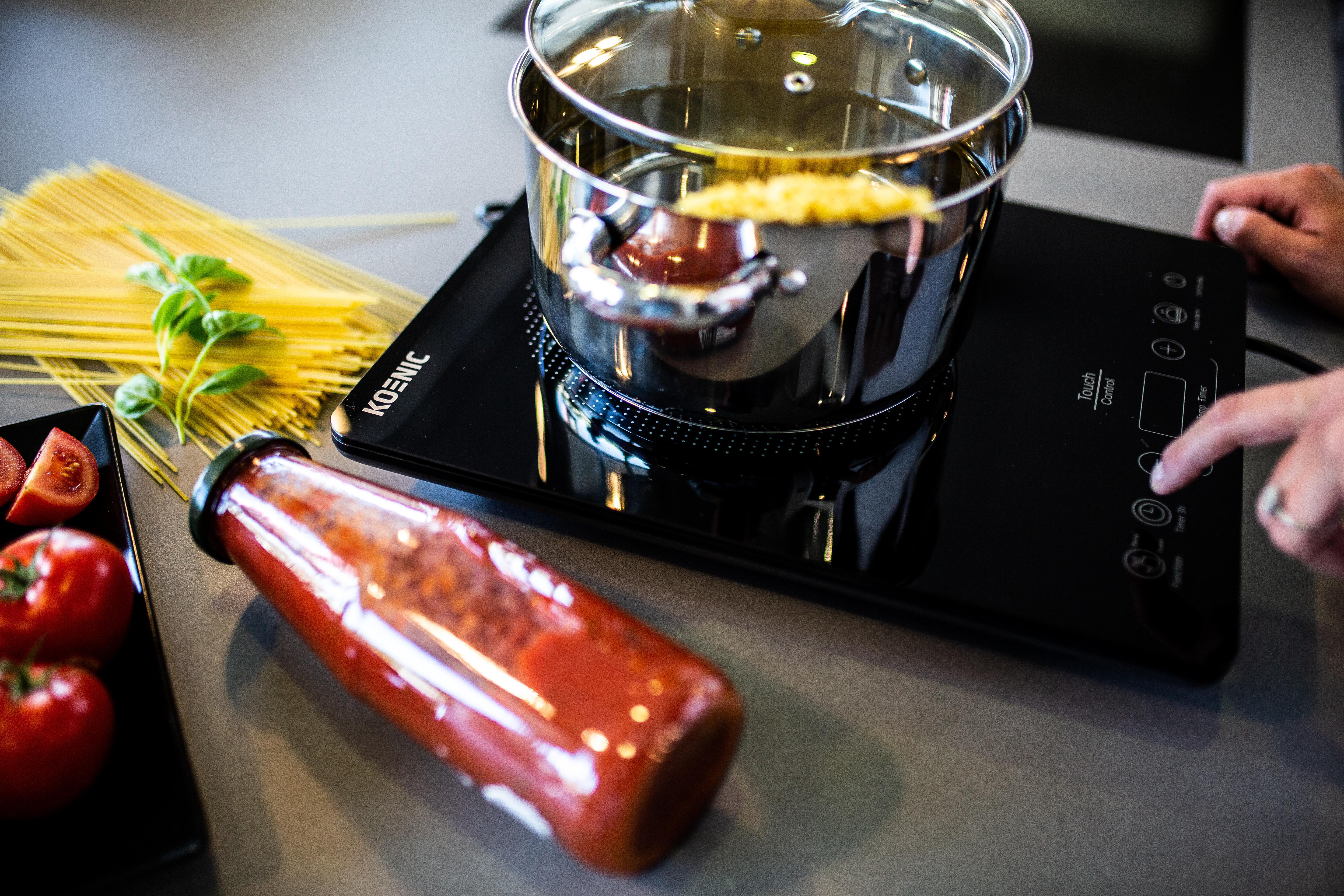 Kochszene mit Topf, Wasser, Nudeln, Tomatensauce und Zutaten auf schwarzem Herd.