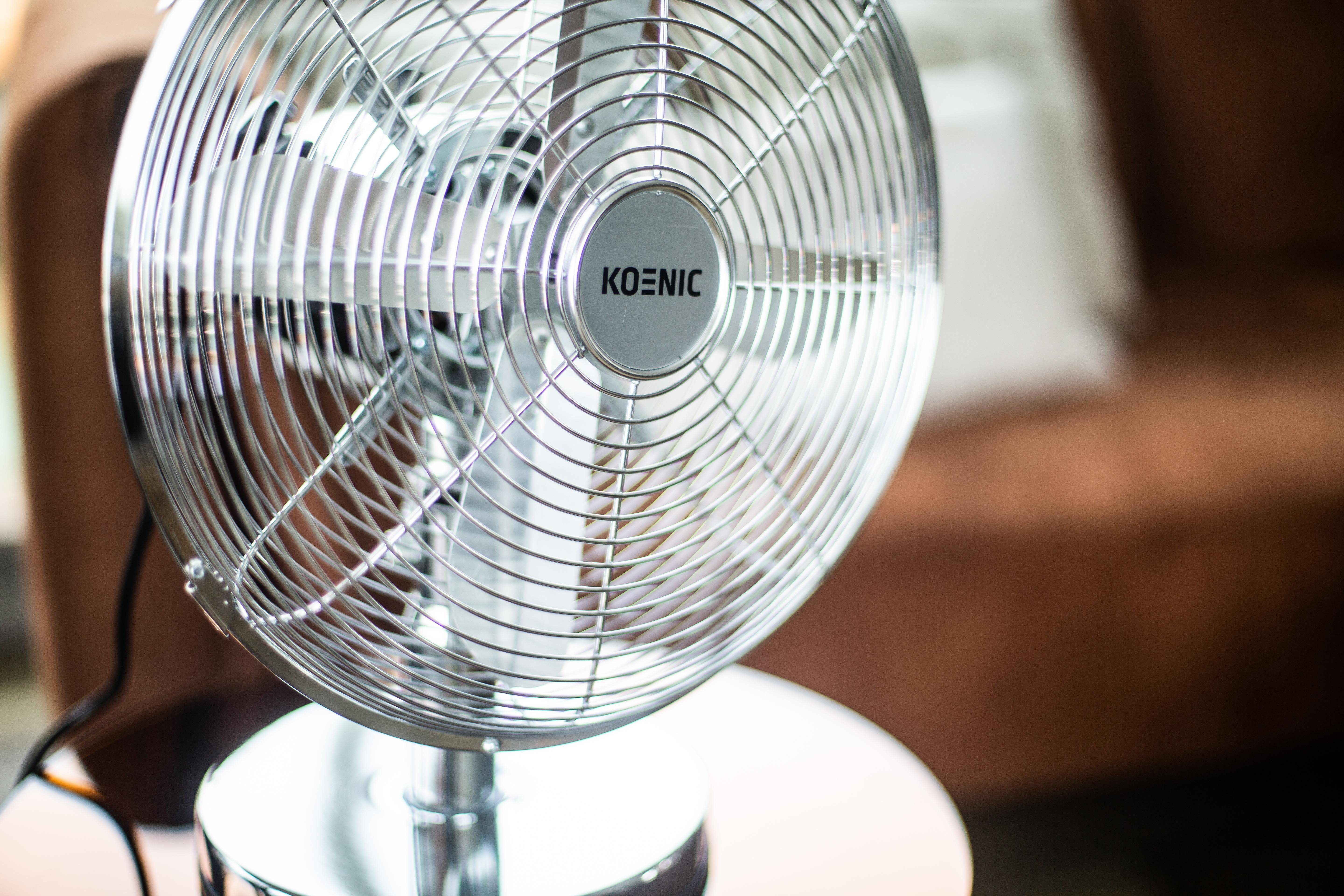 Ein silberner Koenic-Ventilator auf einem weißen Tisch, braunes Sofa im Hintergrund.