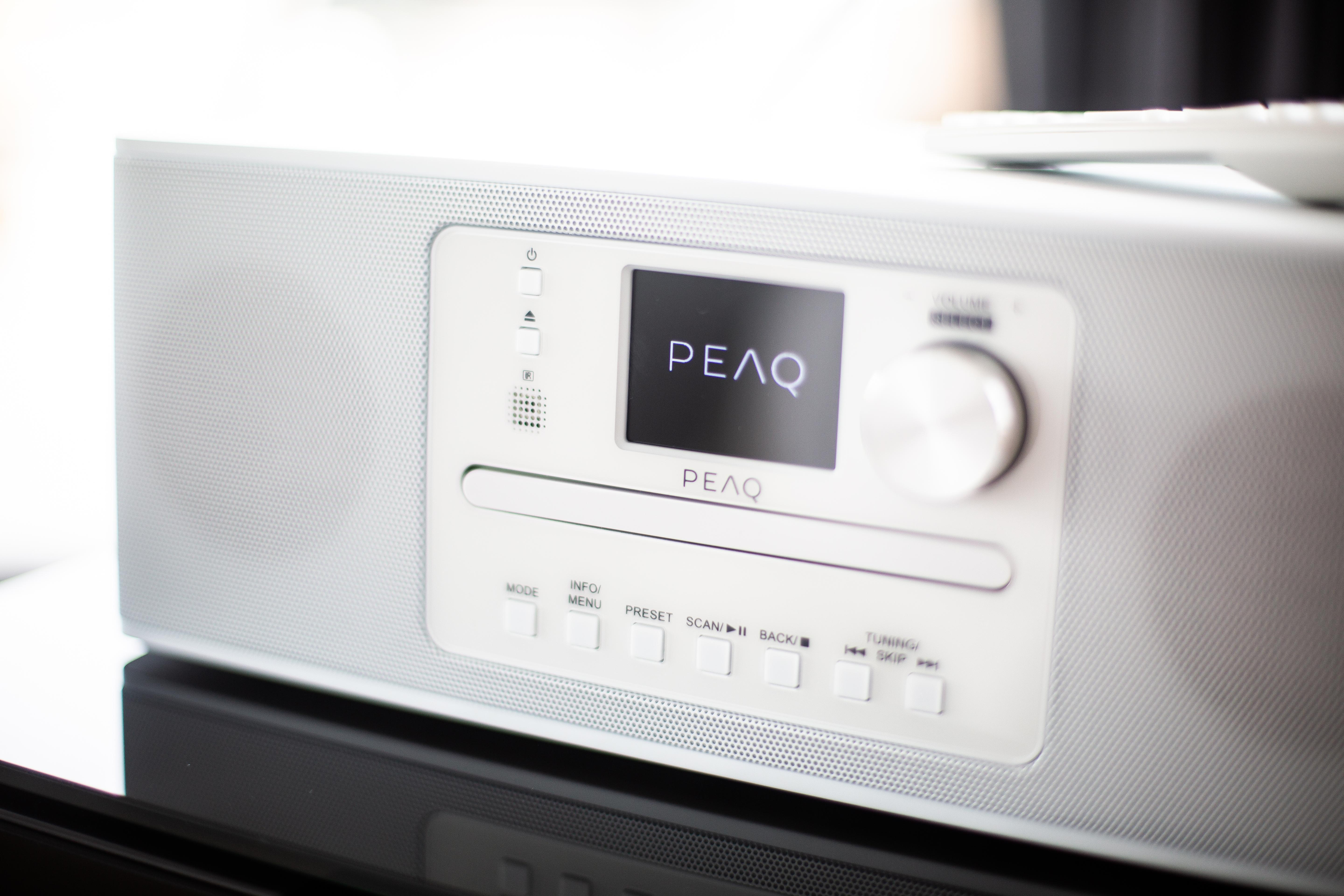 Ein weißes Audiosystem mit einem Display, auf dem 'PEAQ' steht. Knöpfe und ein Drehregler sind sichtbar.