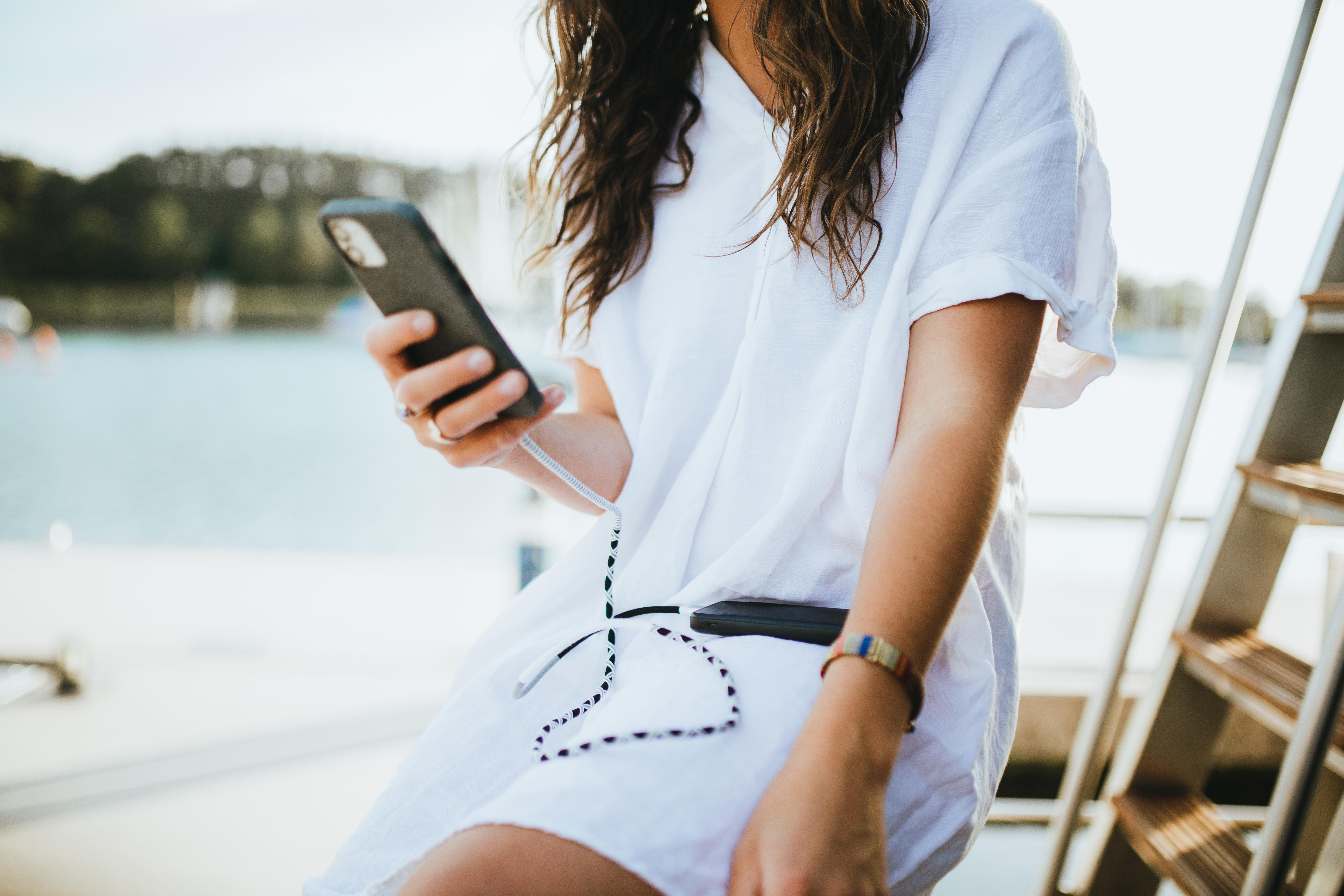 Eine Frau in einem weißen Kleid hält ein Telefon mit Ladekabel in der Nähe eines Bootes.