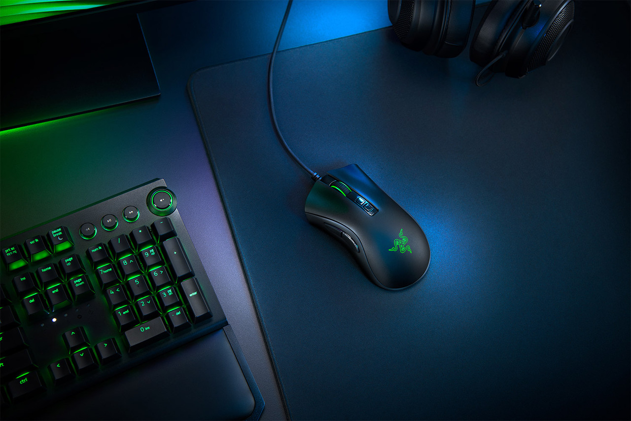 Black gaming mouse on a blue mousepad with a keyboard. Green lights are visible.