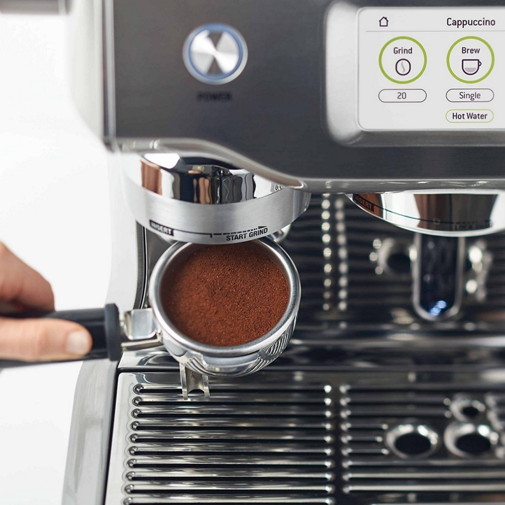 Gros plan sur une machine à expresso. Une main tient un porte-filtre rempli de café moulu. L'écran affiche des options.