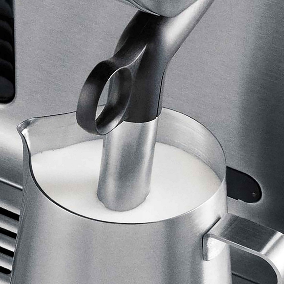 Une baguette à mousser est dans un pichet en acier inoxydable rempli de lait. Le pichet est sur une machine à café. Le lait est blanc.
