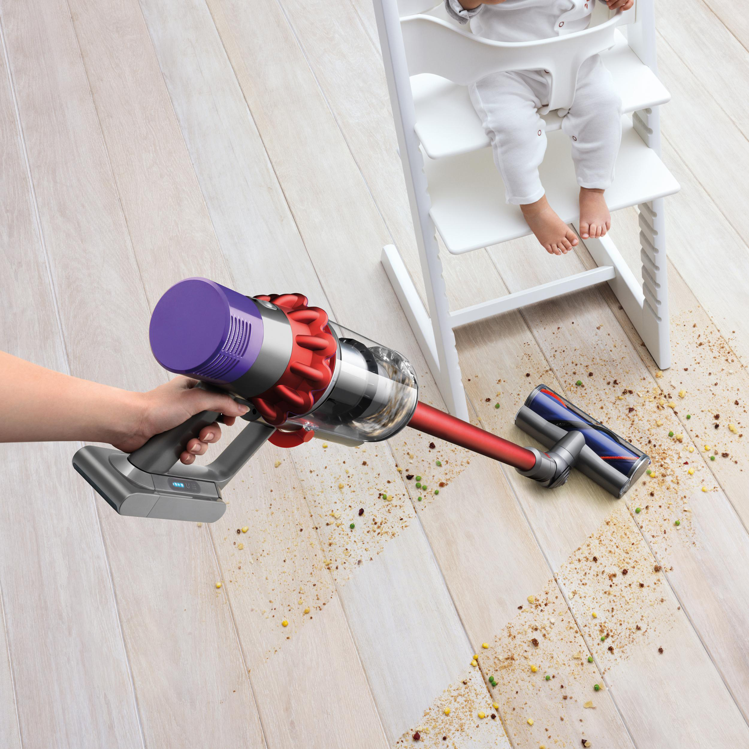 Eine Person saugt einen Holzboden mit einem roten und grauen Dyson-Staubsauger in der Nähe eines Babys im Hochstuhl.