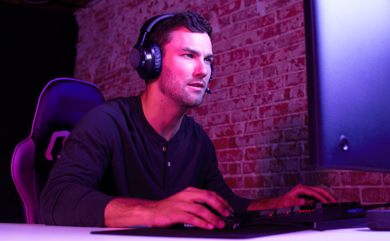 Ein Mann mit Kopfhörern spielt Videospiele. Er ist in einem Raum mit roten Ziegeln, violett beleuchtet.