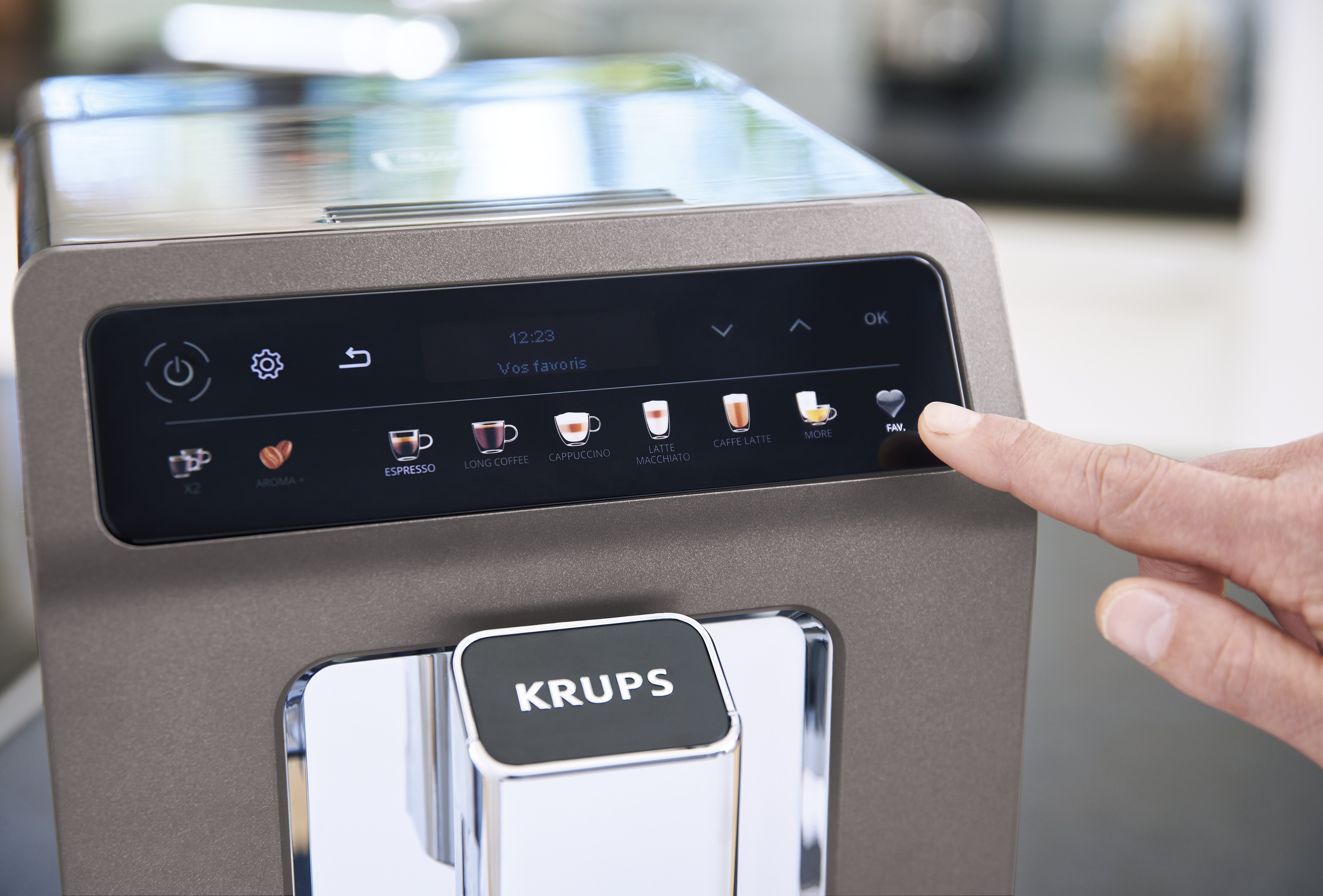 Közelkép egy Krups kávéfőzőről, egy ujj megnyomja a 'Milk' gombot.