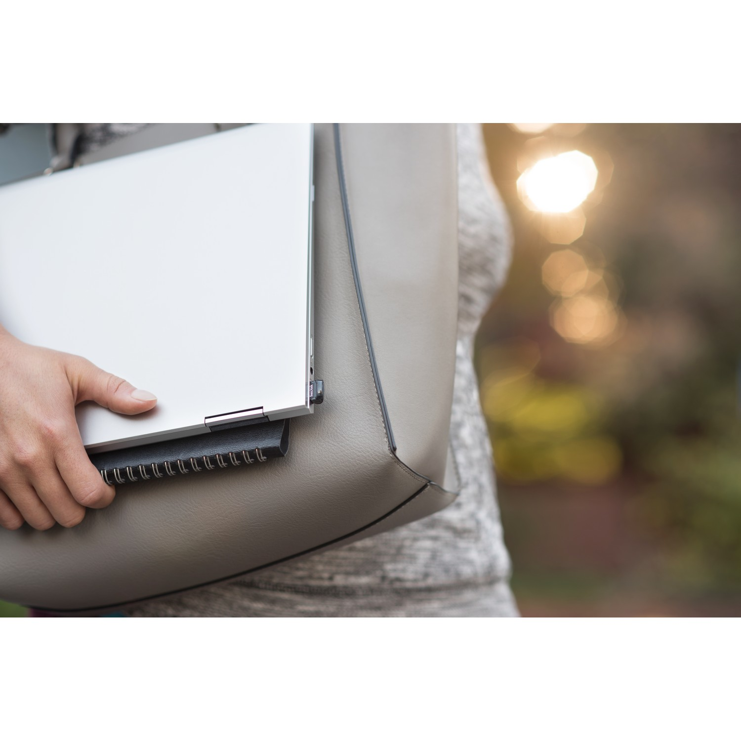 Una persona sosteniendo una laptop y un cuaderno en una bolsa gris, con un fondo borroso al aire libre.