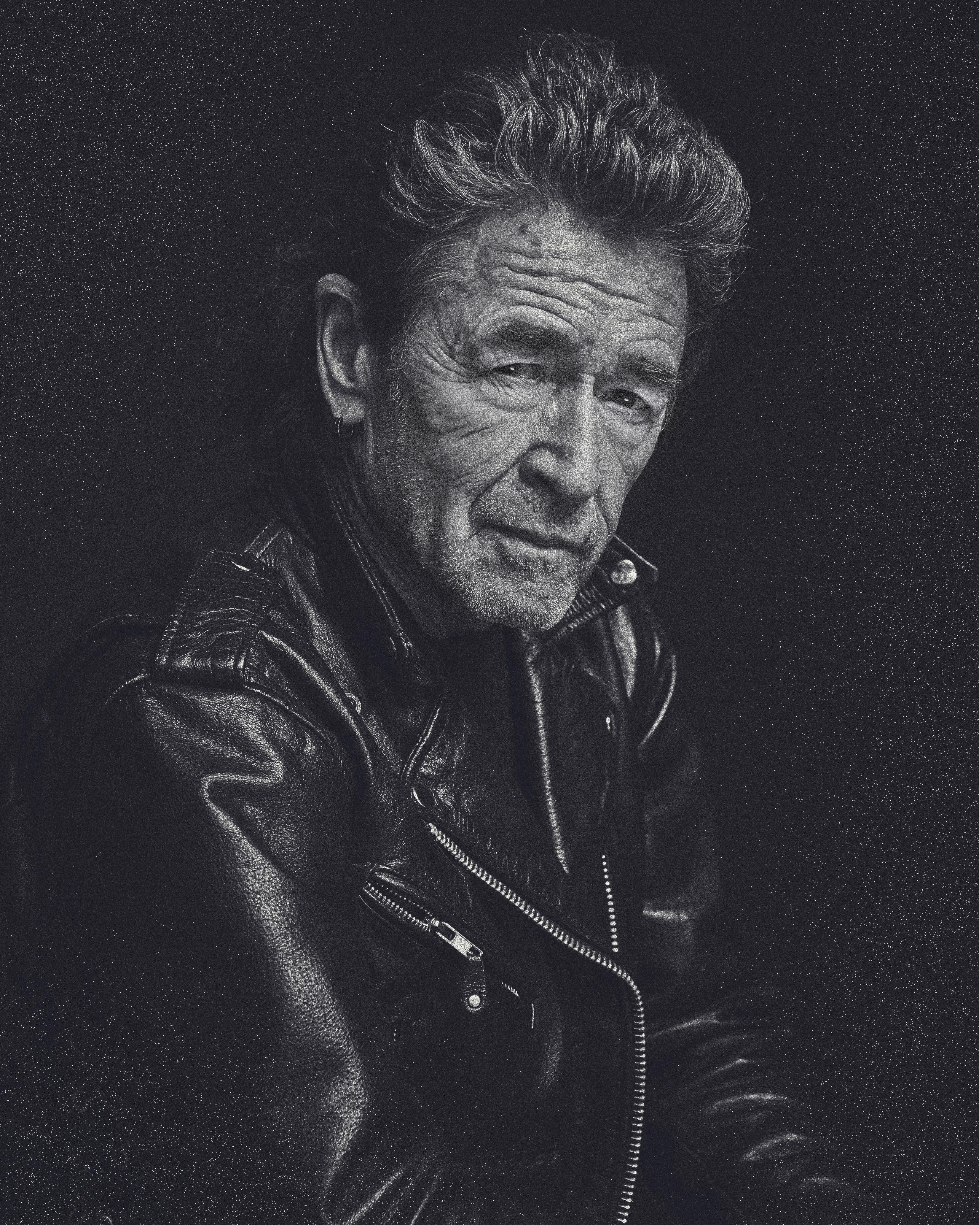 Ein Schwarzweißfoto eines älteren Mannes in einer Lederjacke, der in die Kamera schaut.