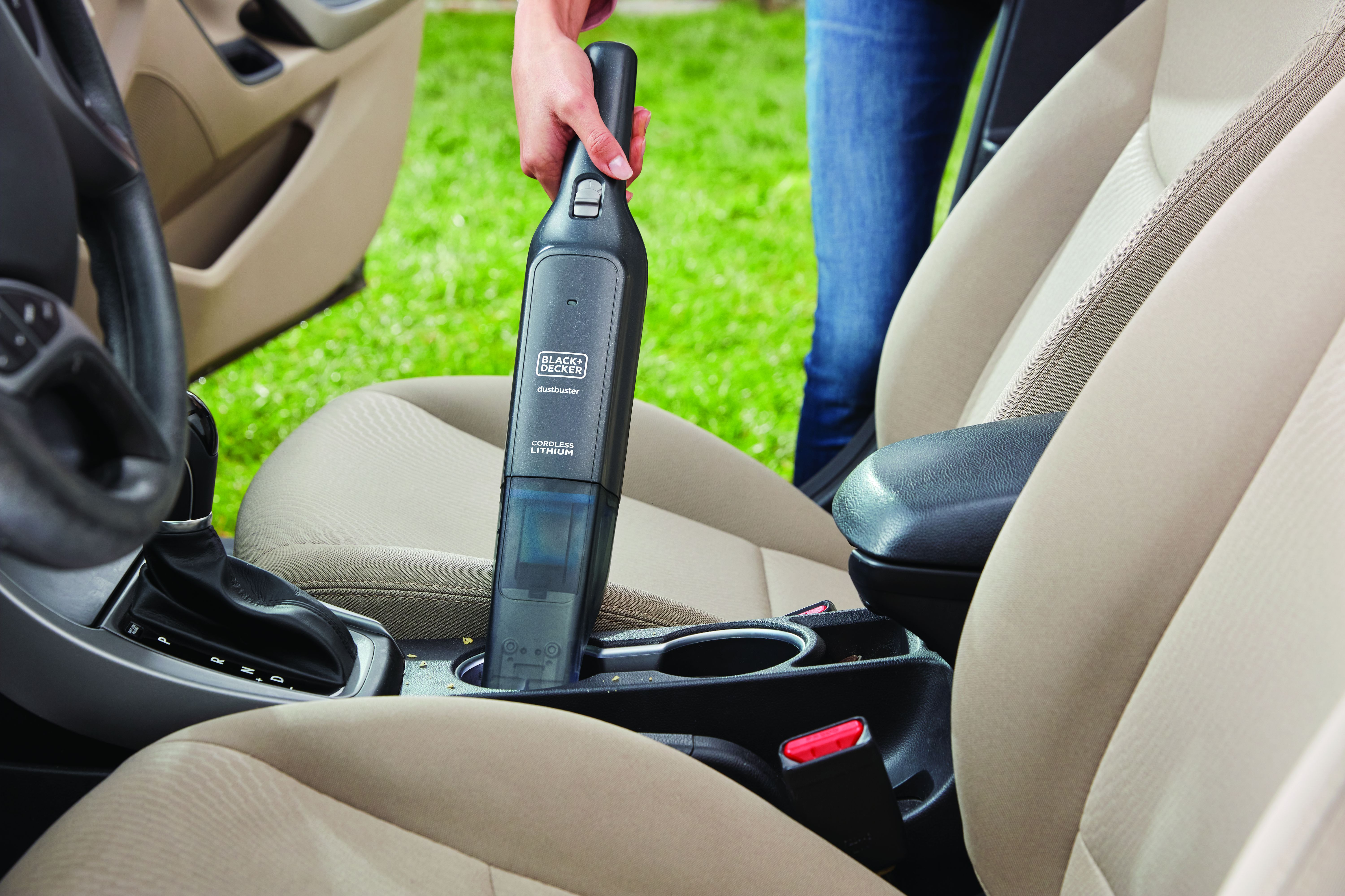 Eine Hand hält einen schwarzen Autostaubsauger in einem Auto. Beige Sitze und ein grüner Rasen sind im Hintergrund.