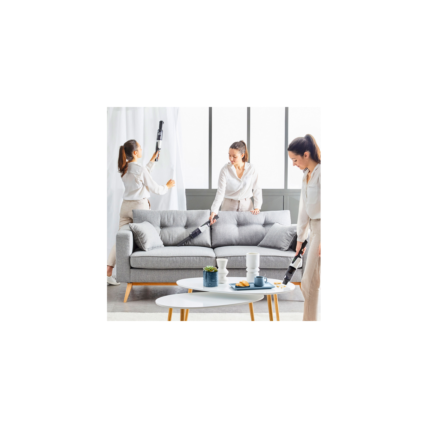 Tres mujeres limpiando una habitación con aspiradoras, sofá gris y mesas blancas presentes.