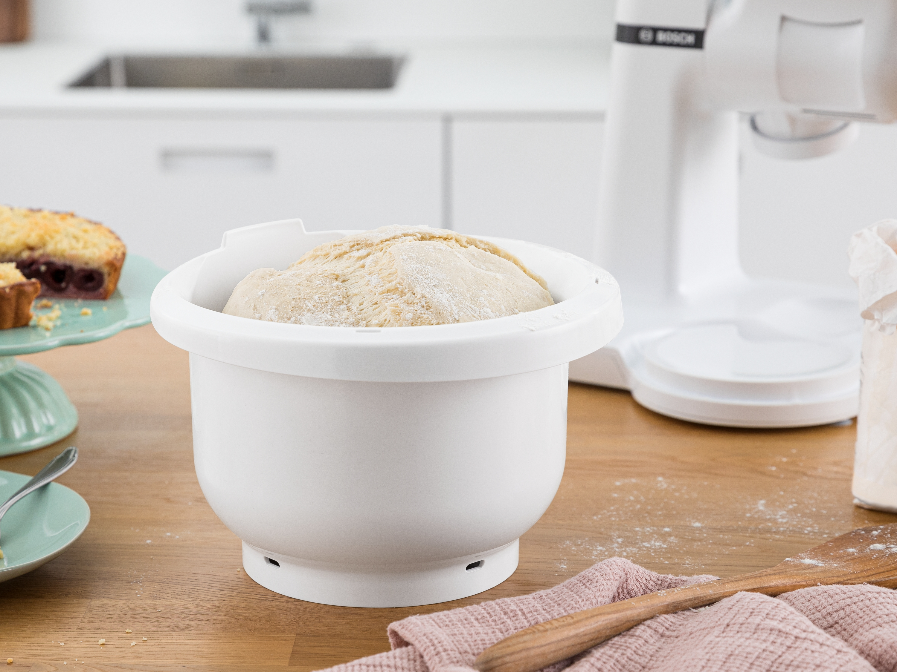 Eine weiße Mixerschüssel mit Teig auf einem Holztisch, mit Backutensilien im Hintergrund.