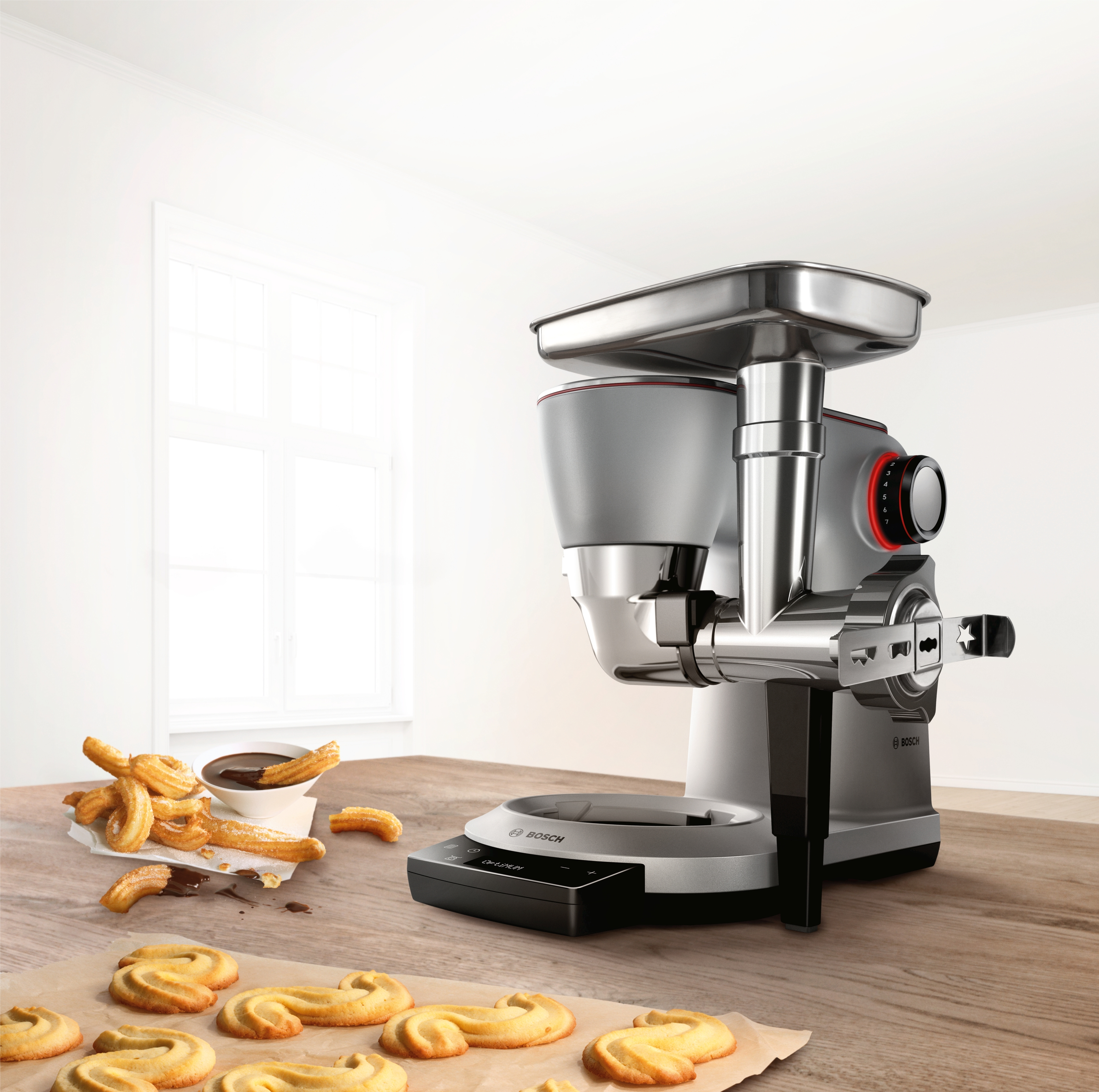 Eine Küchenmaschine mit Keksen und Churros auf einem Holztisch in der Nähe eines Fensters.