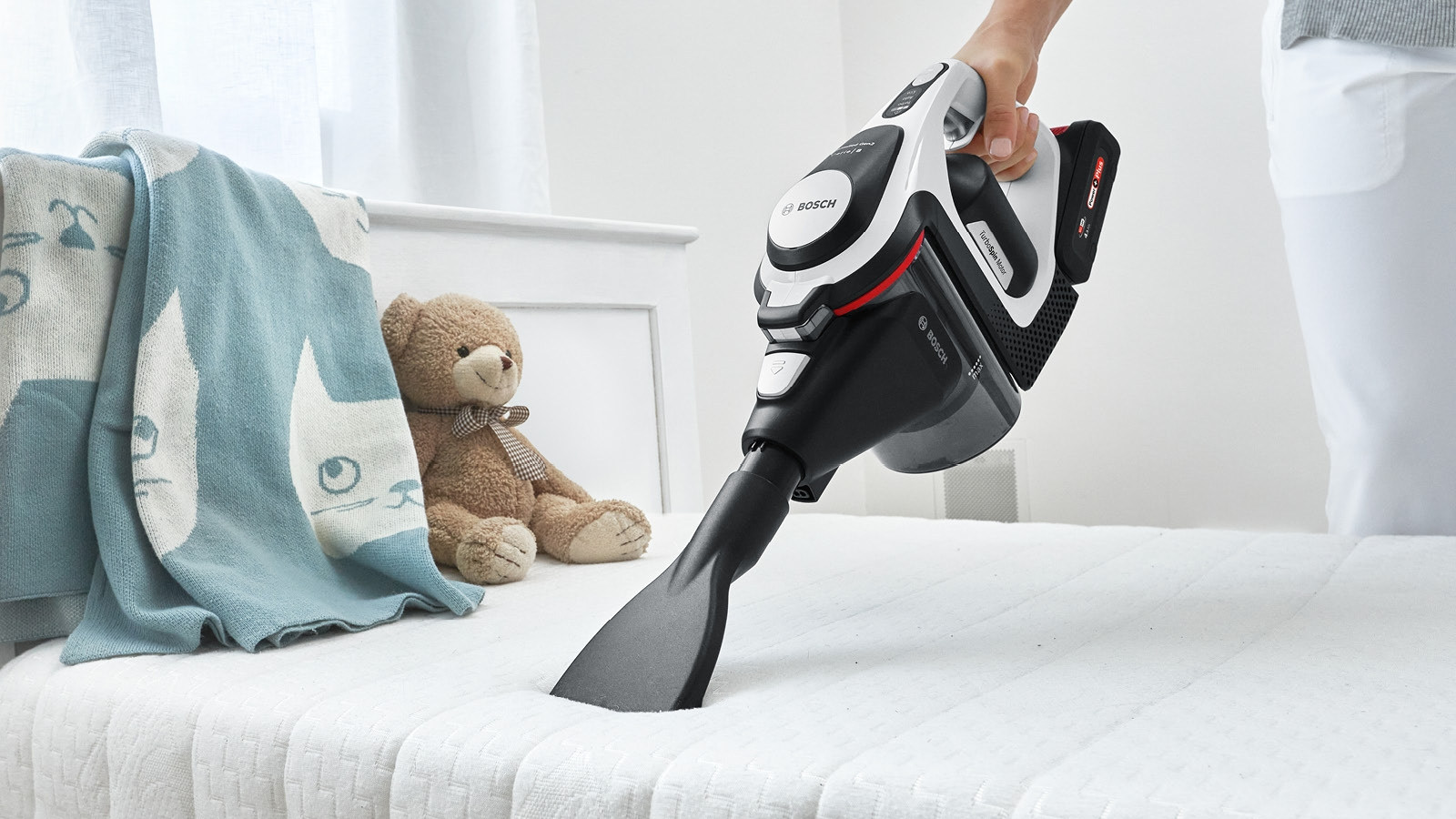 Eine Person saugt ein weißes Bett mit einem schwarz-weißen Bosch-Staubsauger. Eine Decke mit Katzen und ein Teddybär liegen auf dem Bett.