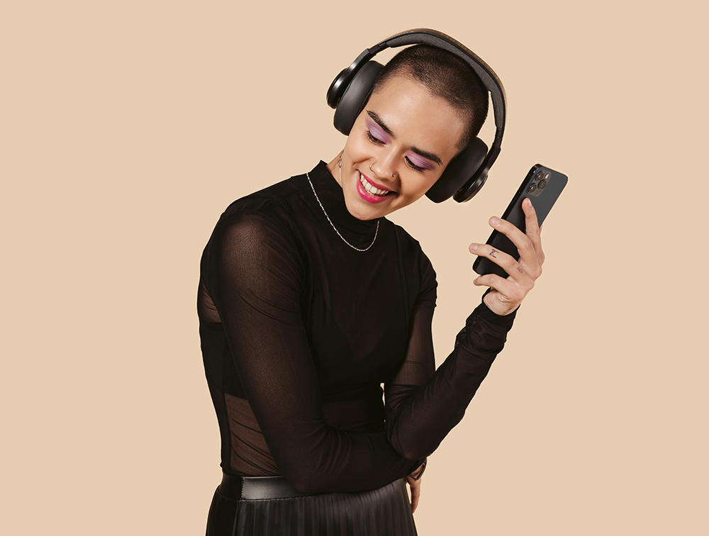 Vrouw met kort haar lacht, draagt een koptelefoon en houdt een telefoon vast, tegen een beige achtergrond.