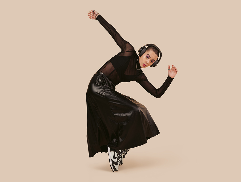 Vrouw in zwarte outfit met koptelefoon, aan het dansen. Beige achtergrond.