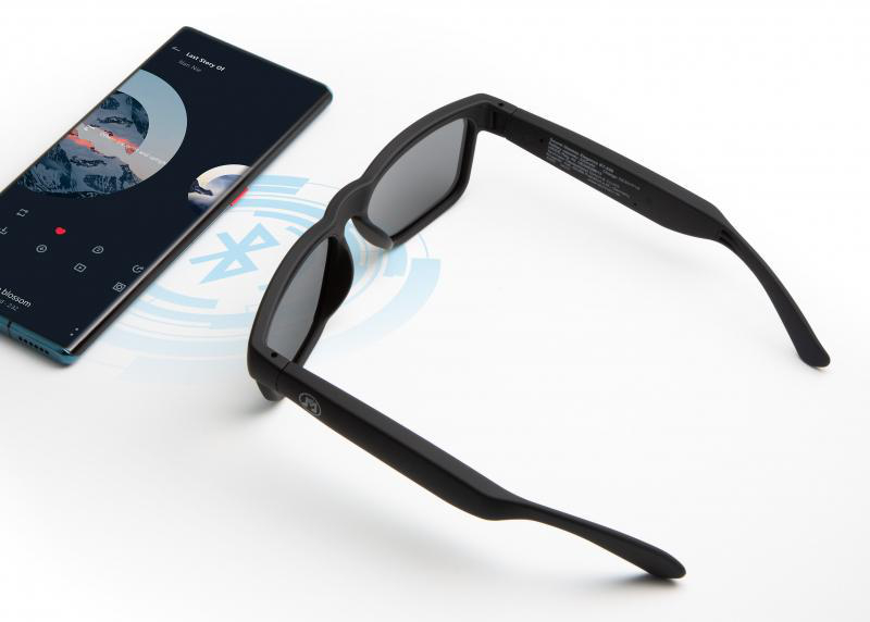 Schwarze Sonnenbrille liegt auf weißer Oberfläche neben einem Telefon mit App.