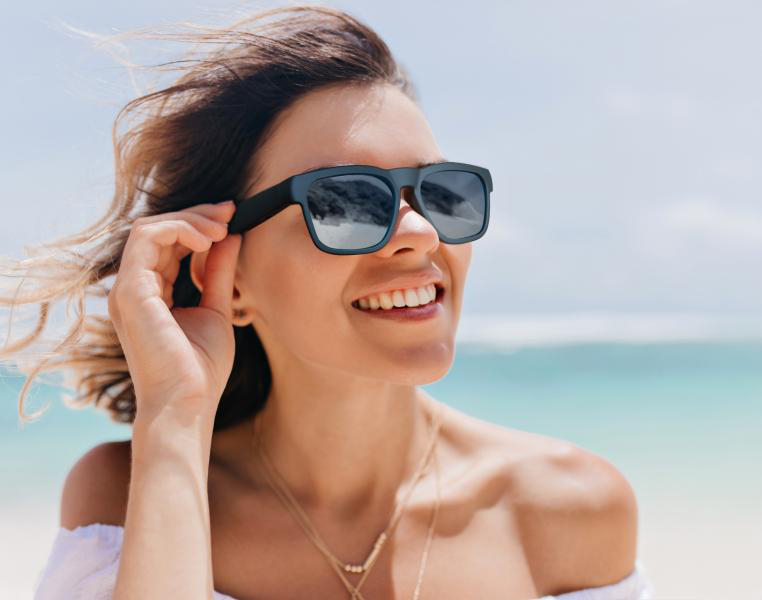 Frau mit Sonnenbrille lächelt am Strand, Hand berührt Brille, sonniger Tag.